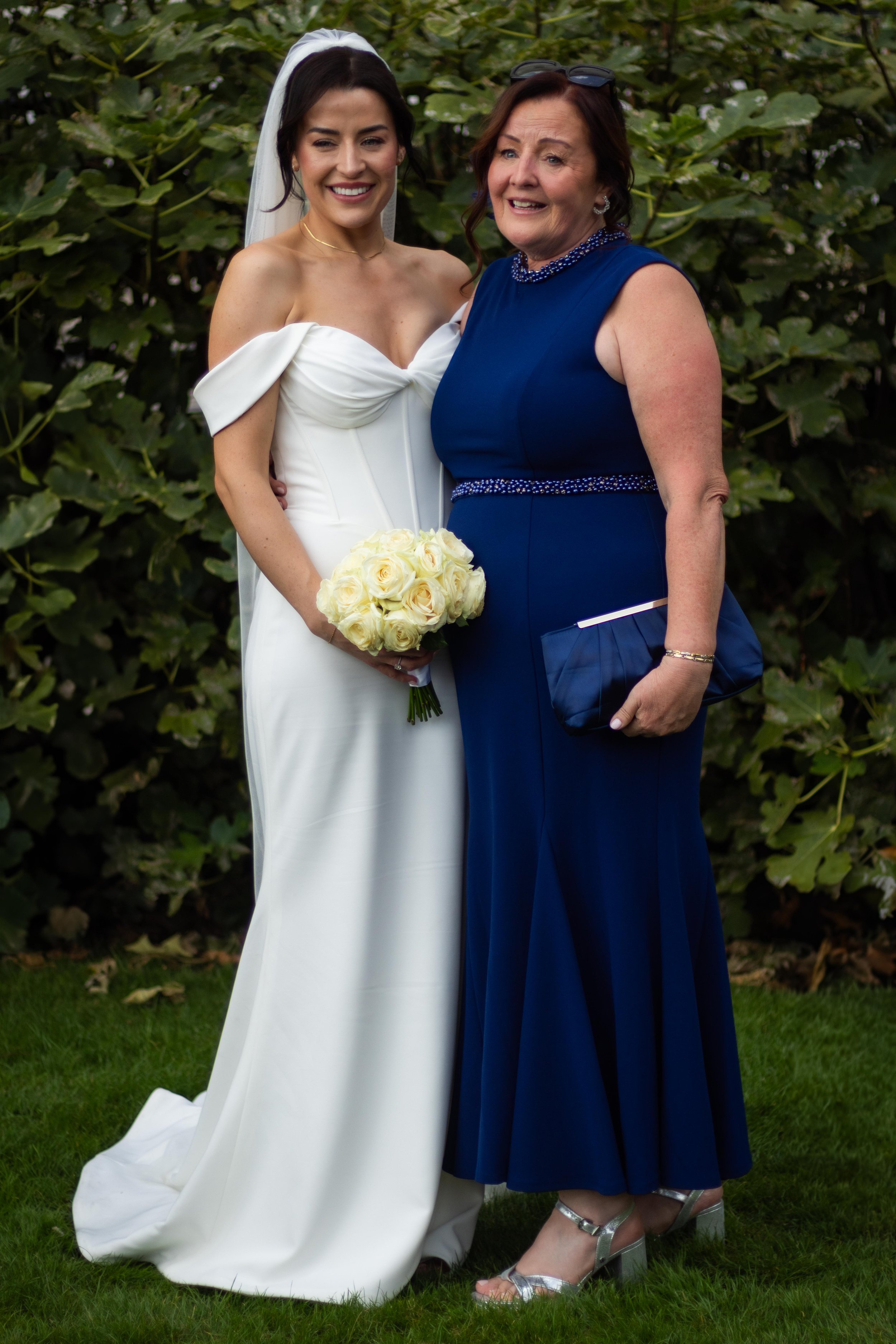 A bride in a white wedding gown holding a bouquet of white roses standing next to a woman in a blue dress with a clutch bag, both smiling outdoors with green foliage in the background.
