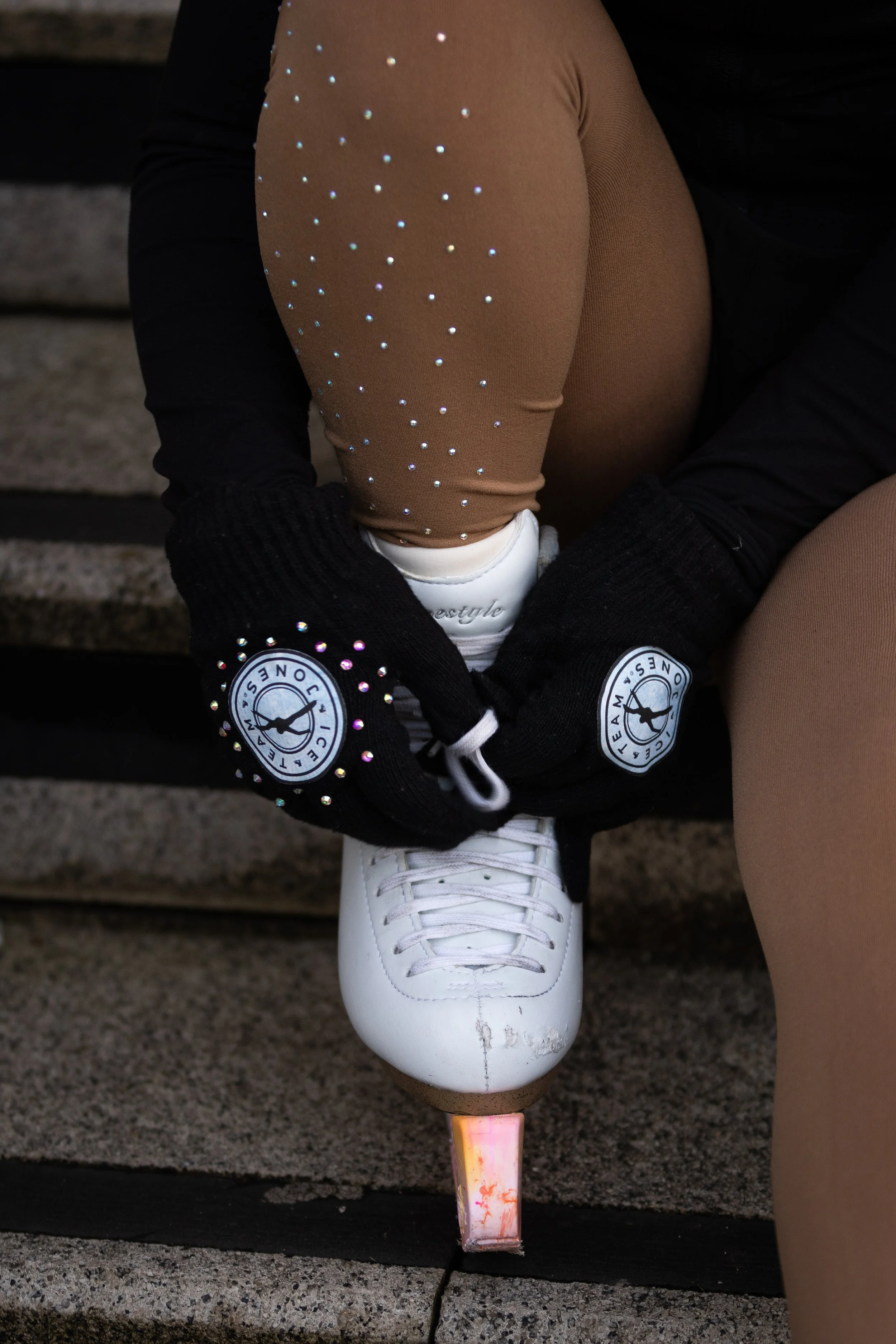 A person wearing beige tights with rhinestone embellishments, black gloves with logo patches, and white figure skates with a pink heel is adjusting their skate on outdoor stone steps.