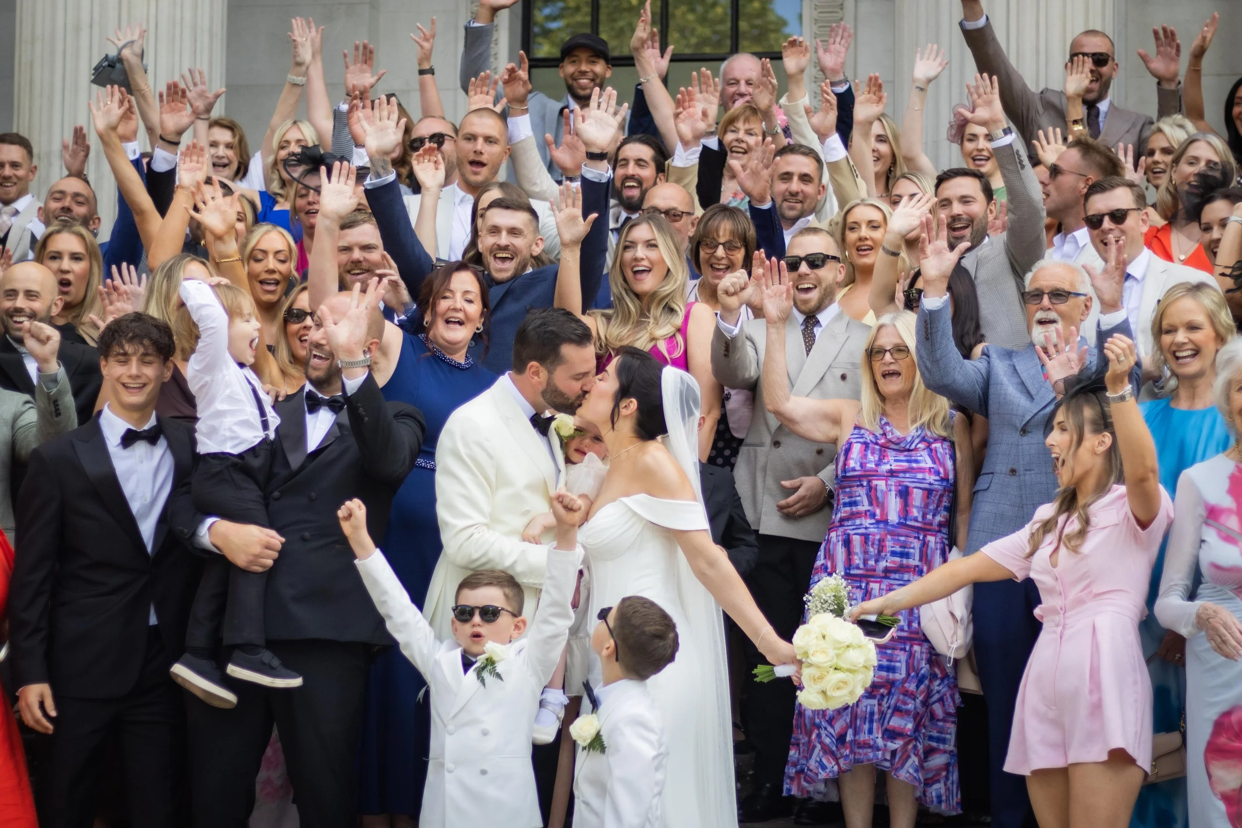 A large group of people celebrating a wedding outside, with a bride and groom kissing in the foreground.