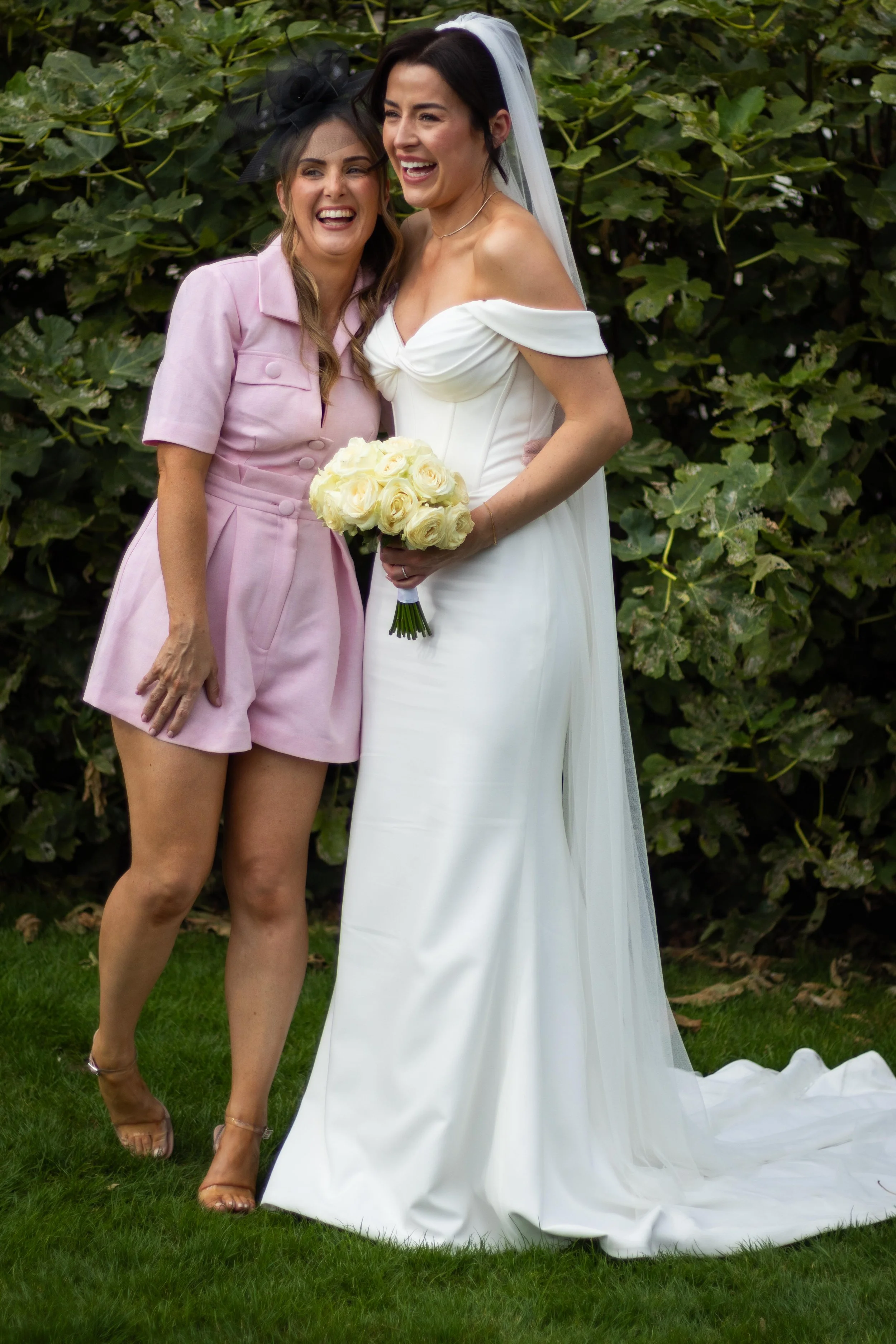 Two women, one in a white wedding dress and the other in a pink outfit, standing together outdoors in front of green foliage, smiling and holding a bouquet of white roses.