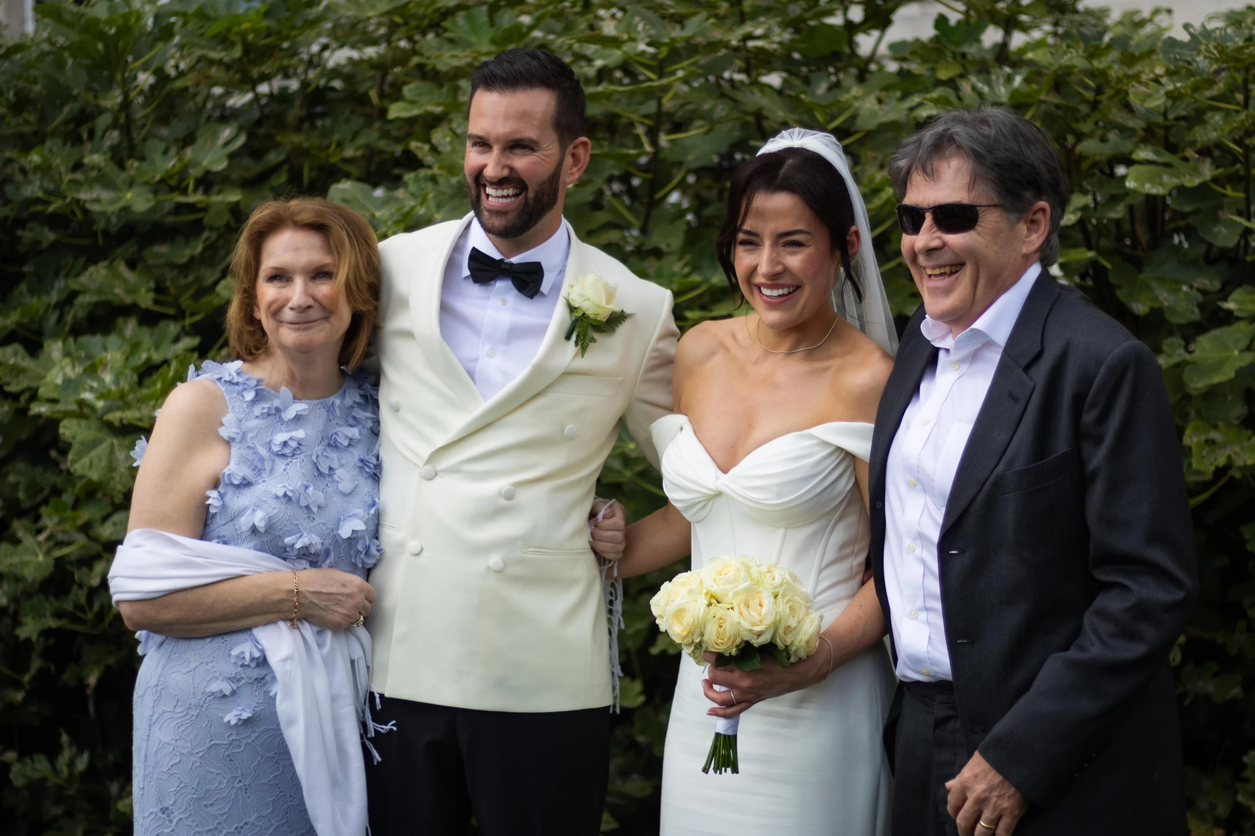 Group of four people smiling at a wedding, with a bride in a white gown holding a bouquet, a groom in a white tuxedo with a black bow tie, and two older adults dressed semi-formally, standing outdoors with greenery in the background.