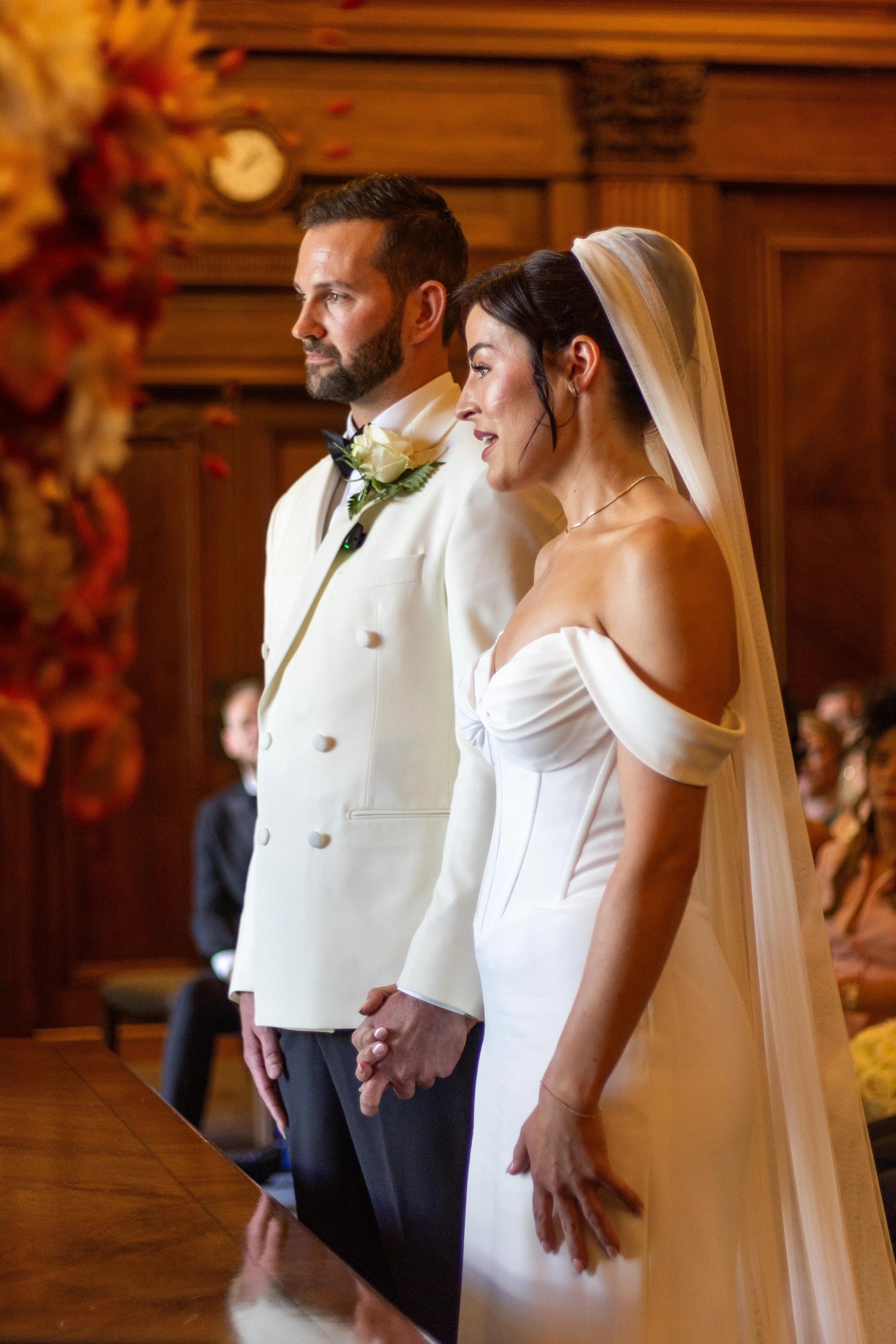 A bride and groom holding hands during their wedding ceremony. The groom is wearing a white tuxedo with a black bow tie and boutonniere. The bride is in a white off-shoulder wedding gown with a veil. They are standing in a wood-paneled room with gues