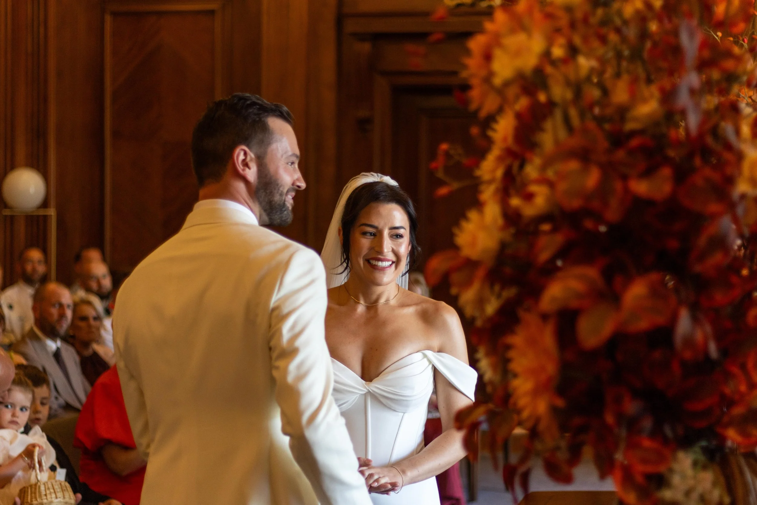 Bride and groom exchanging vows at their wedding ceremony with guests in the background and autumn floral arrangements in the foreground.
