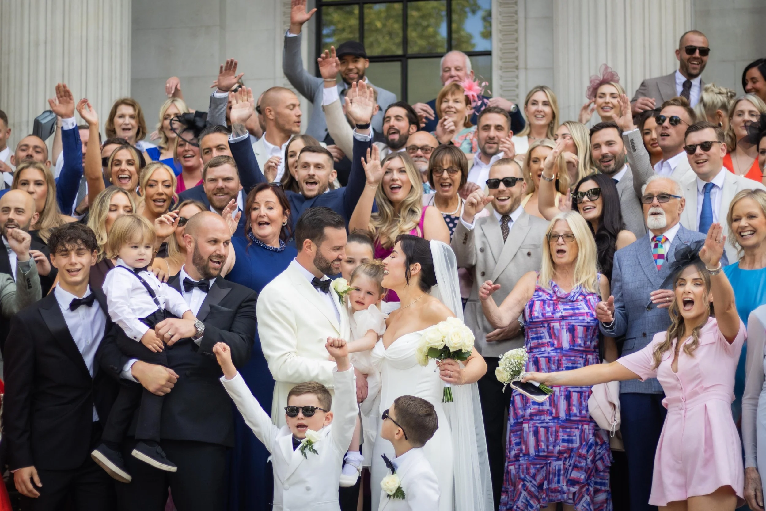 A large group of people celebrating a wedding on the steps of a building, with many raising their hands and smiling. The bride and groom are in the center, holding each other with the bride holding a bouquet of white roses. The guests are dressed in 