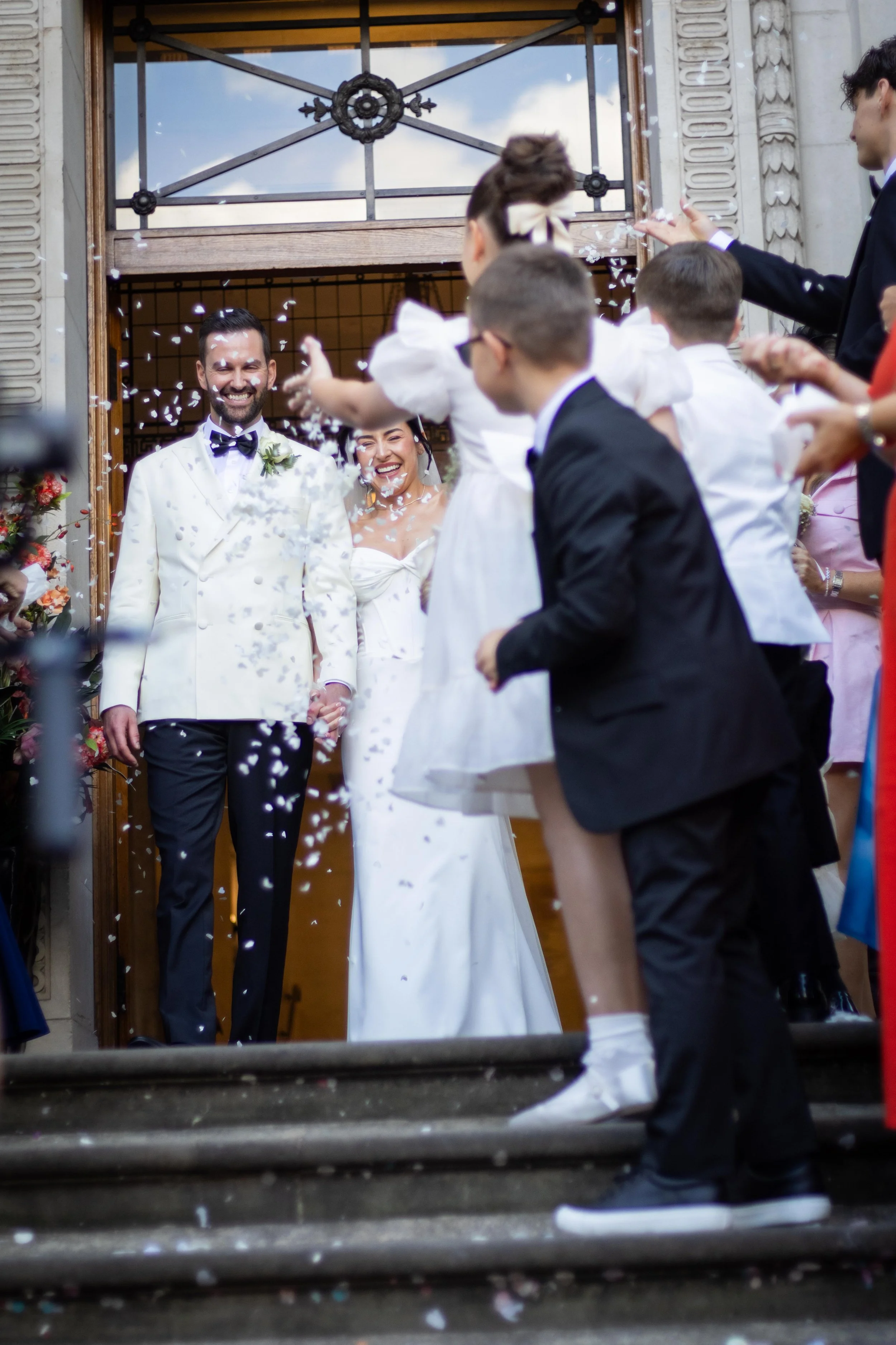 A newlywed couple in wedding attire exiting a building through a door, surrounded by friends and family throwing confetti, celebrating.