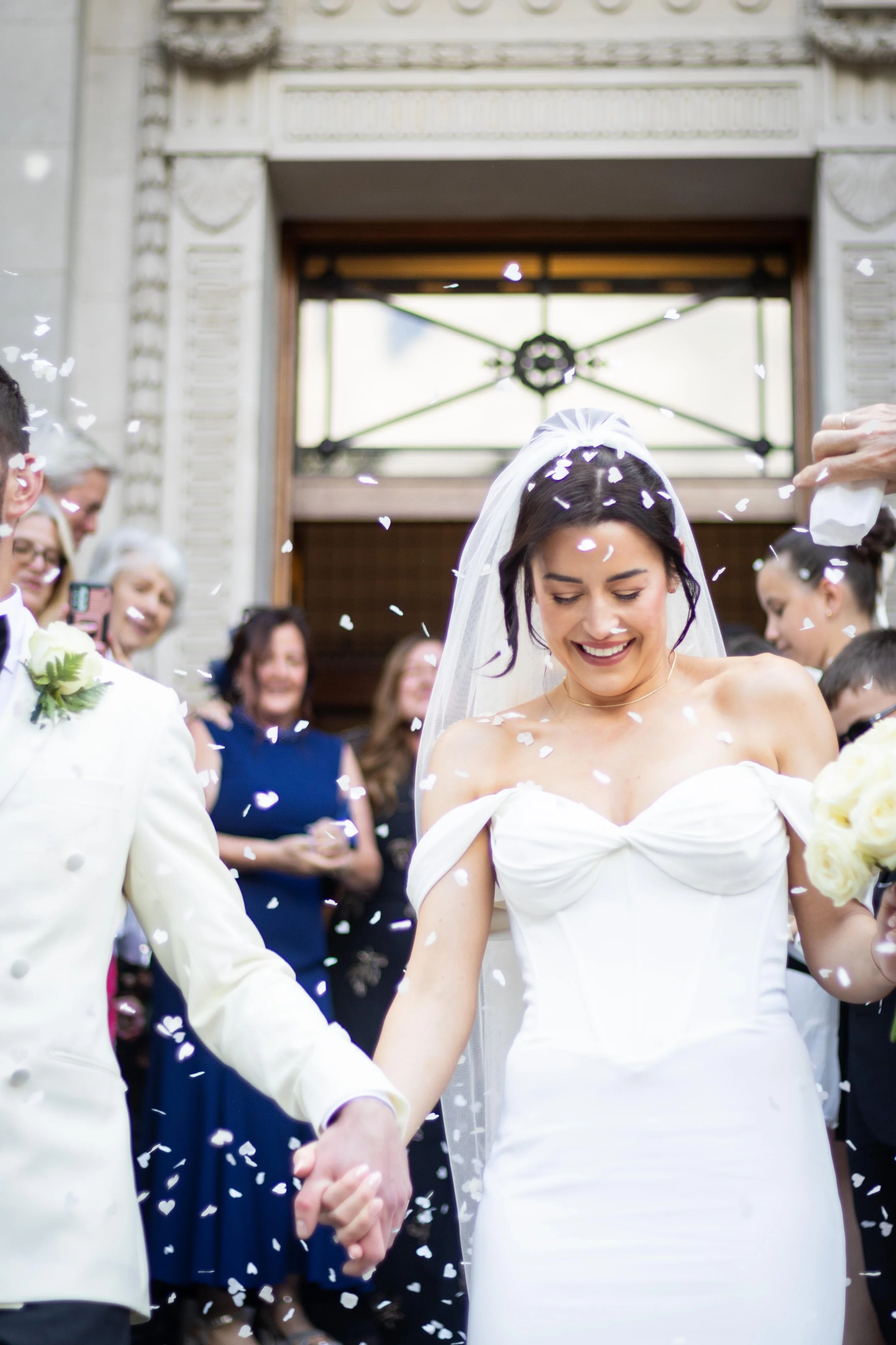 Bride and groom holding hands, smiling, surrounded by guests, as confetti falls at a wedding ceremony.