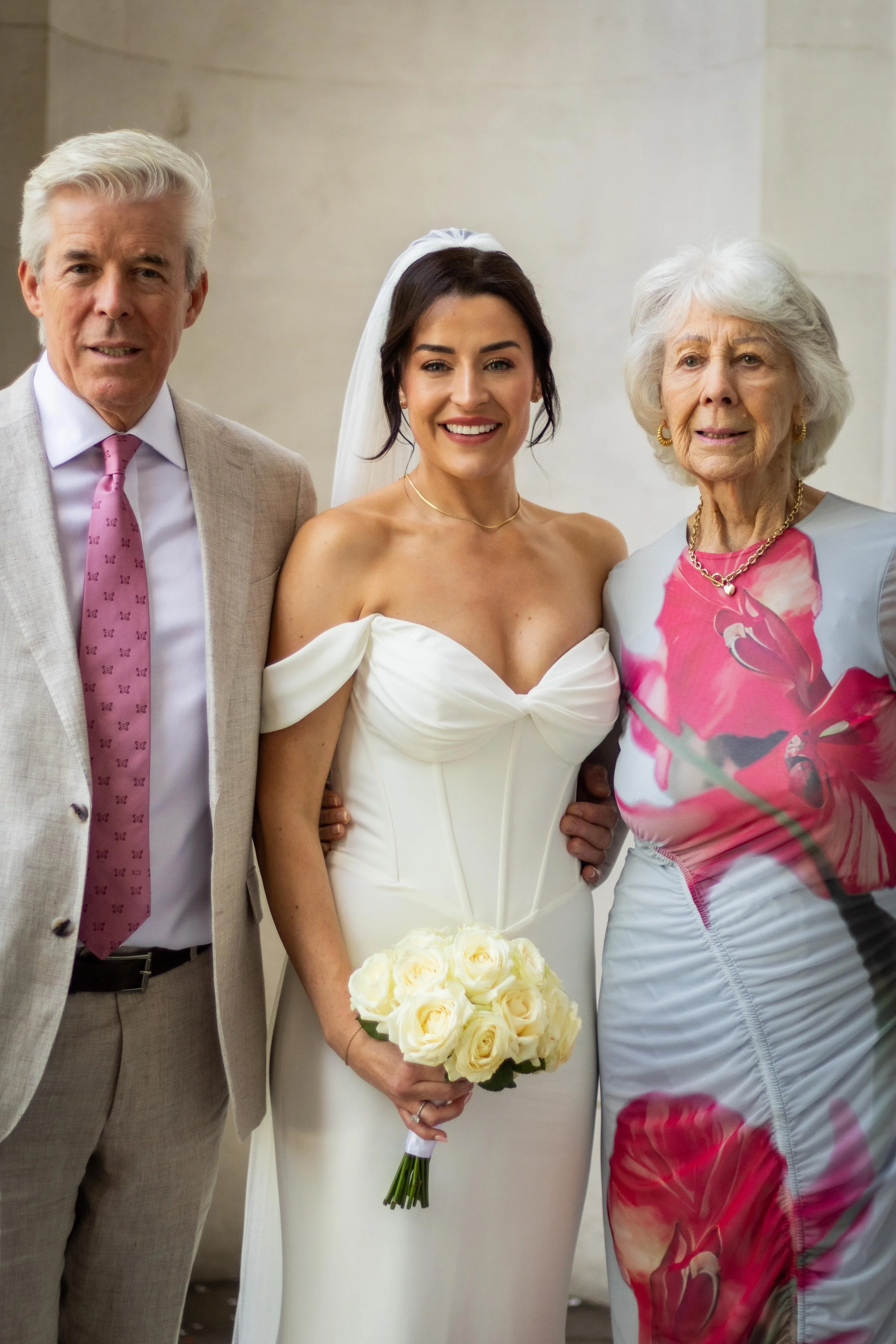 A bride standing between two older women, possibly her parents, at her wedding. The bride wears a white off-the-shoulder wedding gown and holds a bouquet of white roses. The man on her left wears a beige suit with a pink tie, and the woman on her rig