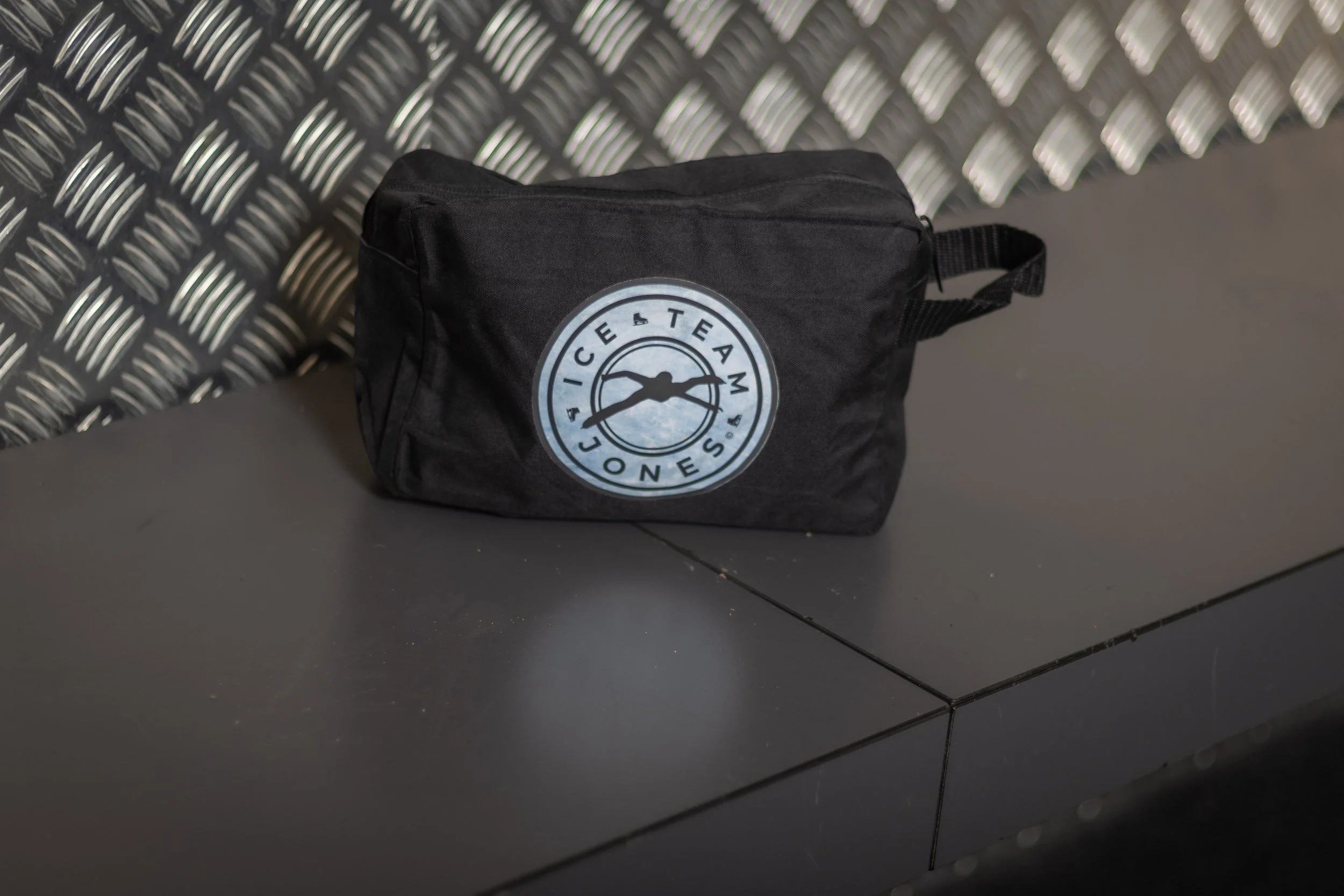 A black bag with a circular logo on it, sitting on a dark gray surface with a metallic textured wall in the background.
