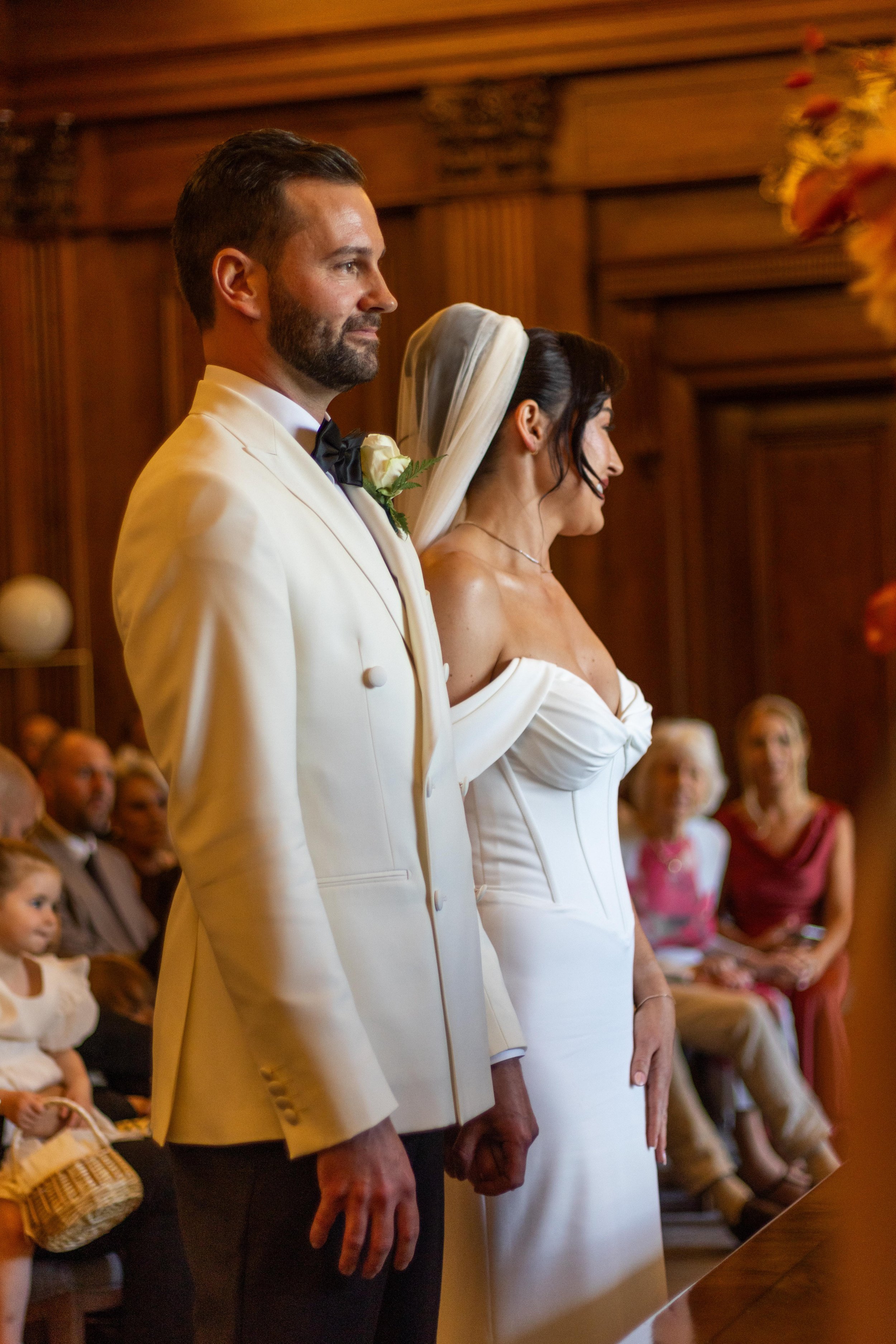 A bride and groom standing side by side during their wedding ceremony, with guests seated in the background in a wood-paneled room.
