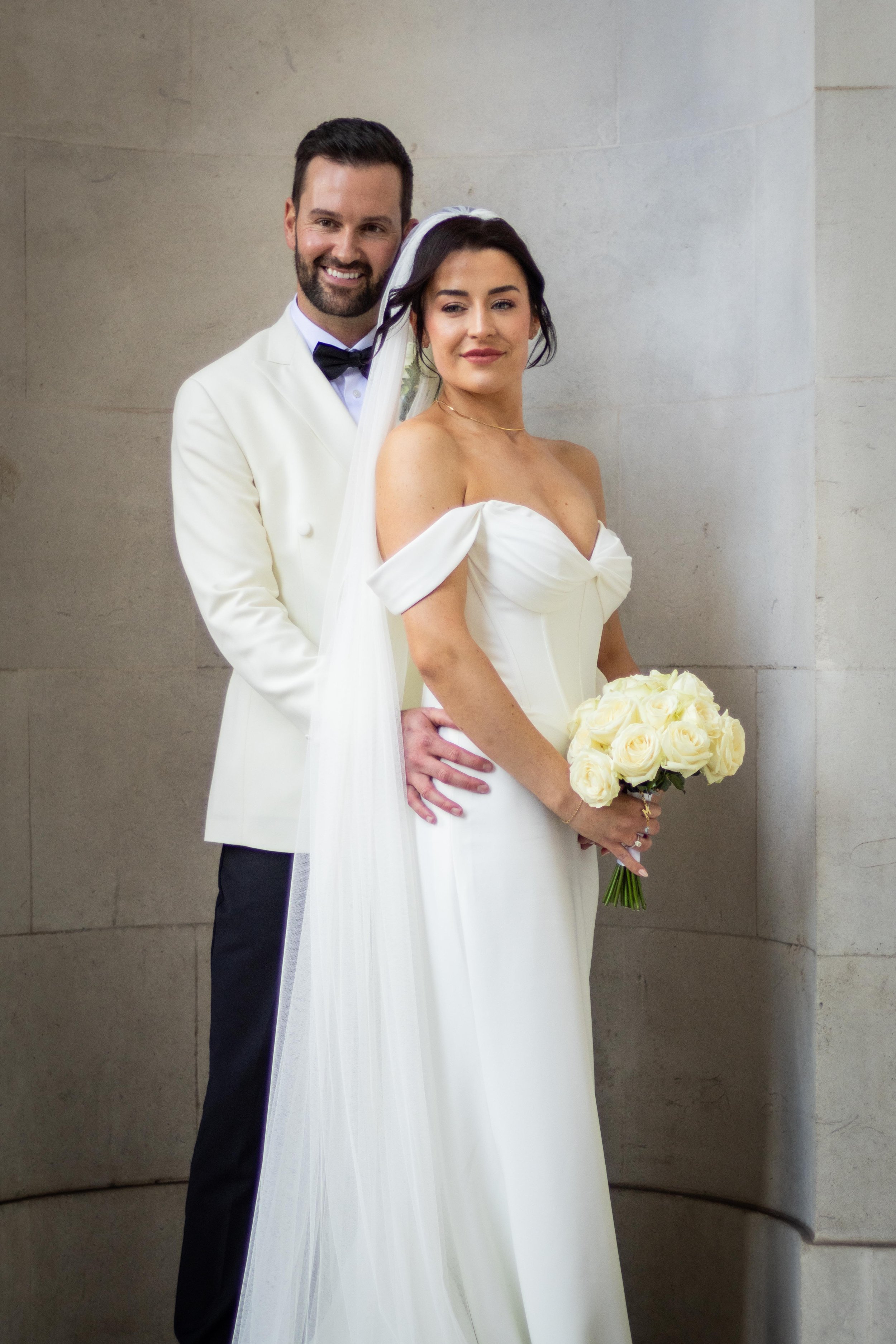 A bride and groom standing together, with the groom embracing the bride from behind, dressed in formal wedding attire. The bride is holding a bouquet of white roses.