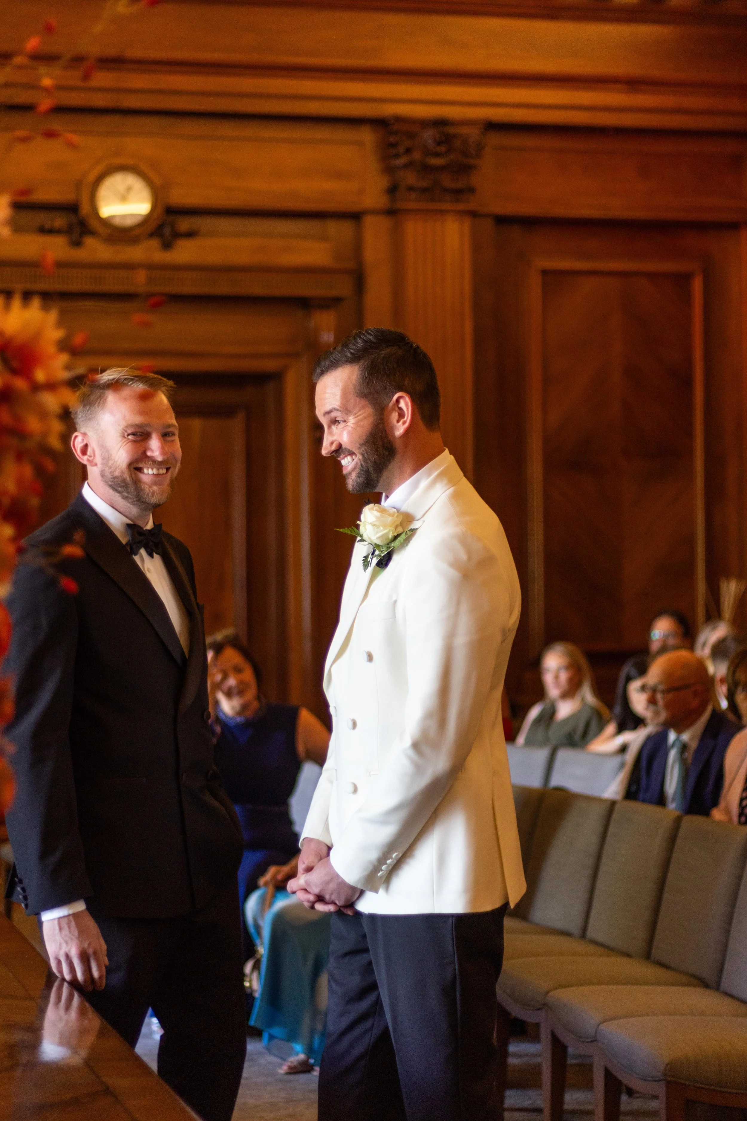 Wedding ceremony with two men, one in a black tuxedo and the other in a white tuxedo, standing in a wood-paneled room with seated guests in the background.