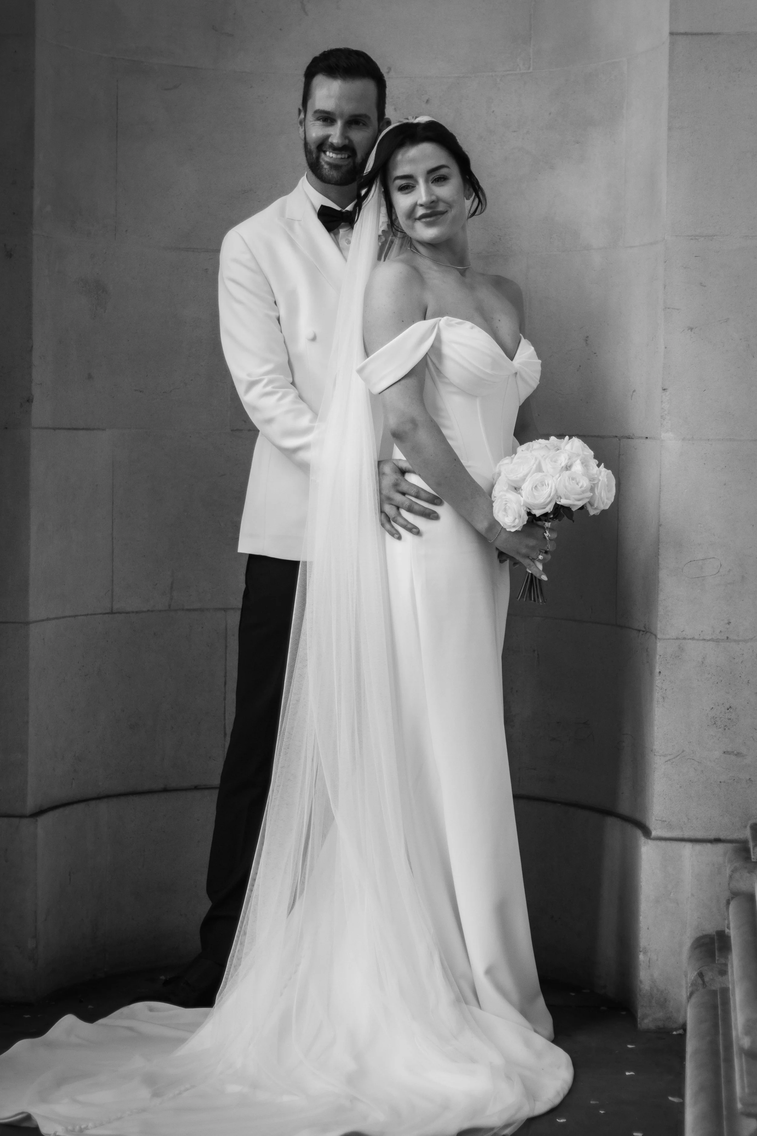 A black and white photo of a bride and groom standing together, with the groom’s arm around the bride's waist. The bride is holding a bouquet of roses and wearing a strapless wedding gown with a long train, while the groom is dressed in a white jacke