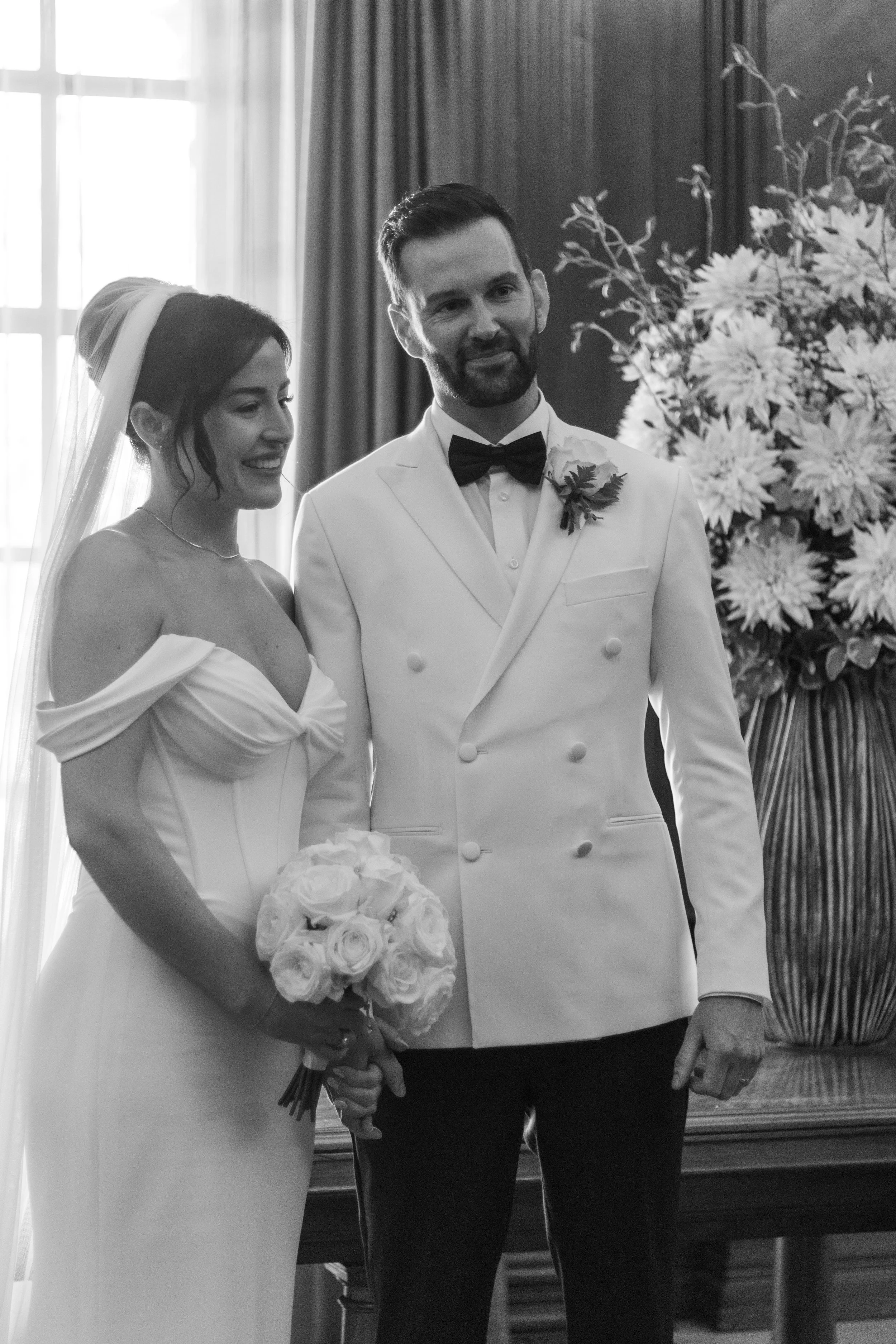 Black and white photo of a bride and groom at their wedding ceremony, with the bride holding a bouquet of roses and the groom wearing a tuxedo with a bow tie, standing in front of a large floral arrangement.