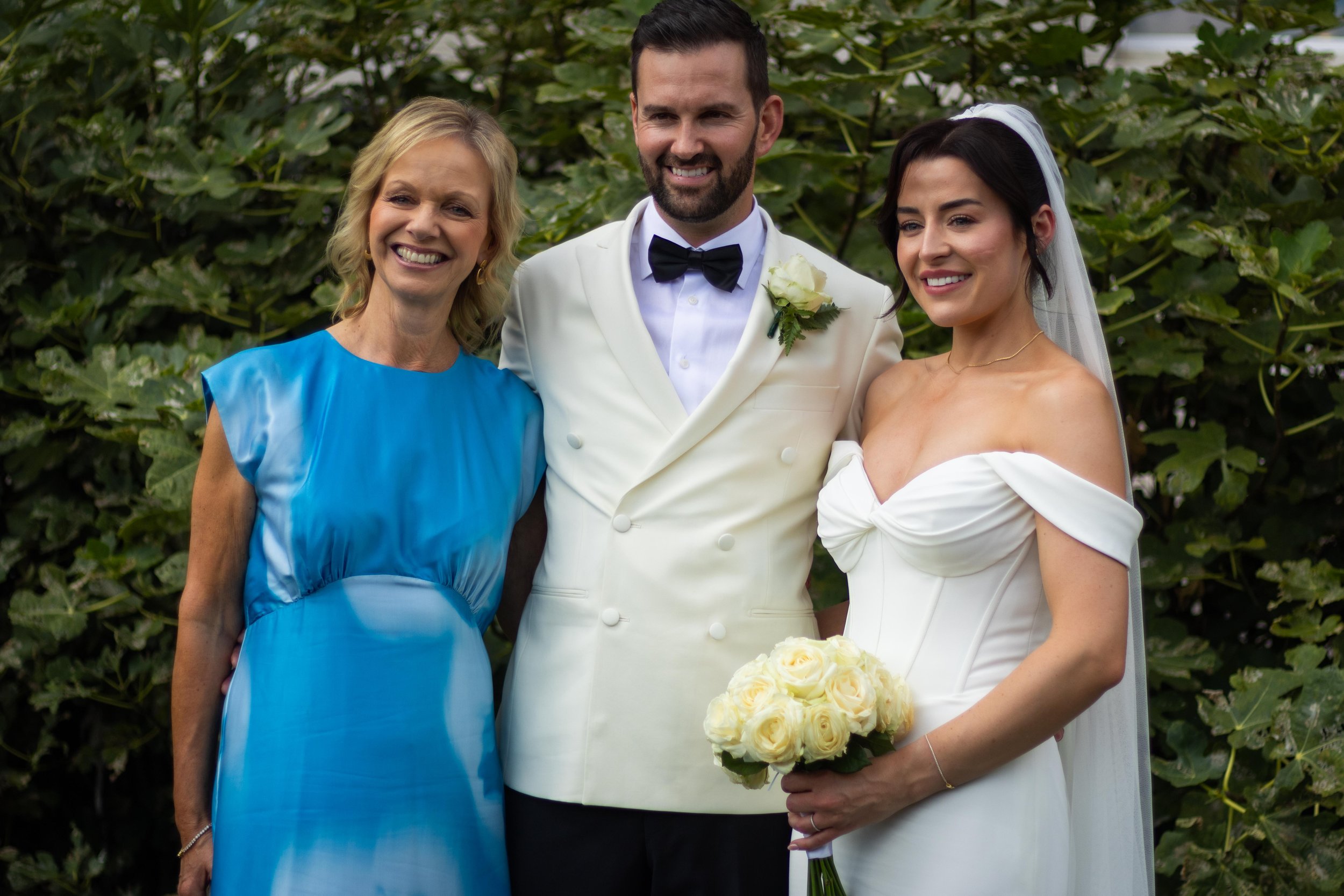 Three people smiling, a woman in a blue dress, a man in a white tuxedo with a black bow tie, and a woman in a wedding gown holding a bouquet of cream roses, standing outdoors with green foliage in the background.