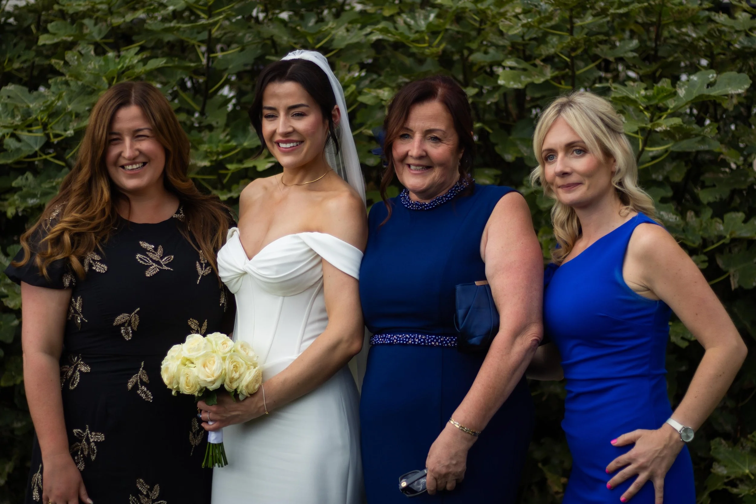 Four women, including a bride in a white wedding dress holding a bouquet of white roses, stand outdoors in front of green foliage, smiling.