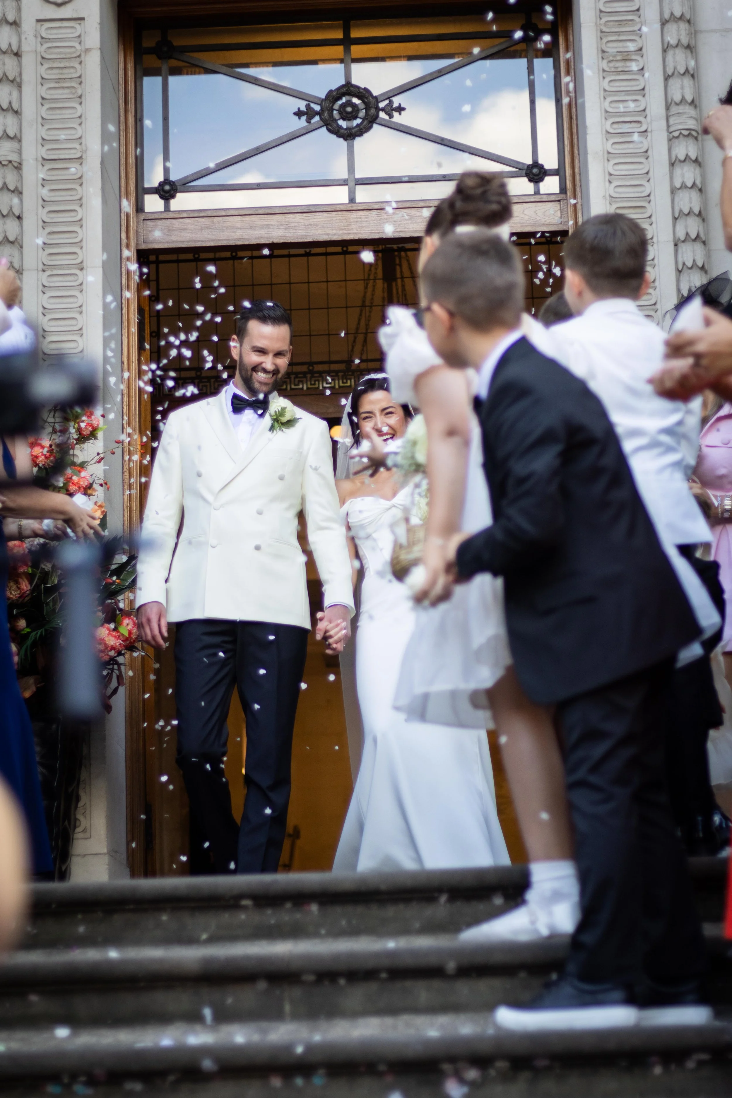A newly married couple holding hands, smiling, as they exit a building and are showered with confetti by friends and family.