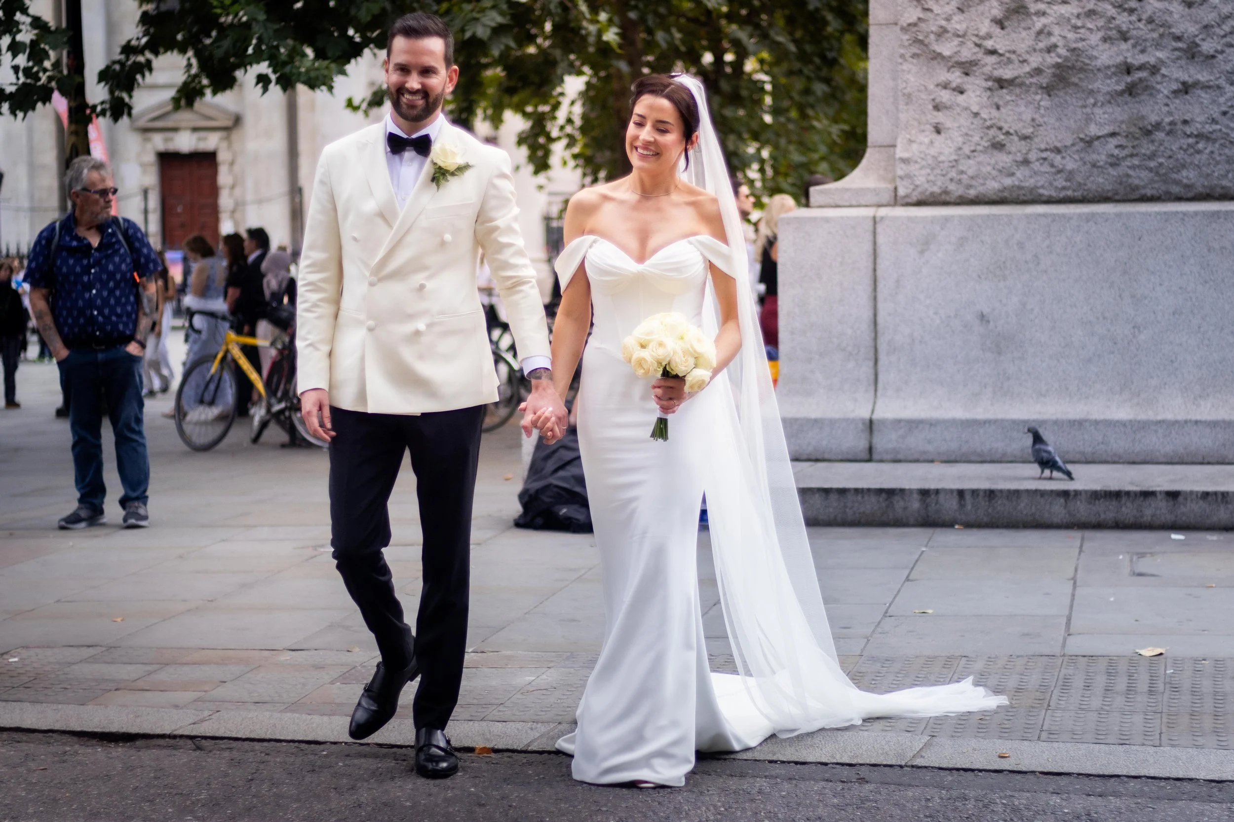 A smiling bride and groom holding hands walking on a city street during daytime, with wedding attire. The bride is wearing a white off-shoulder gown with a veil and holding a bouquet of white roses. The groom is in a white tuxedo jacket with black tr