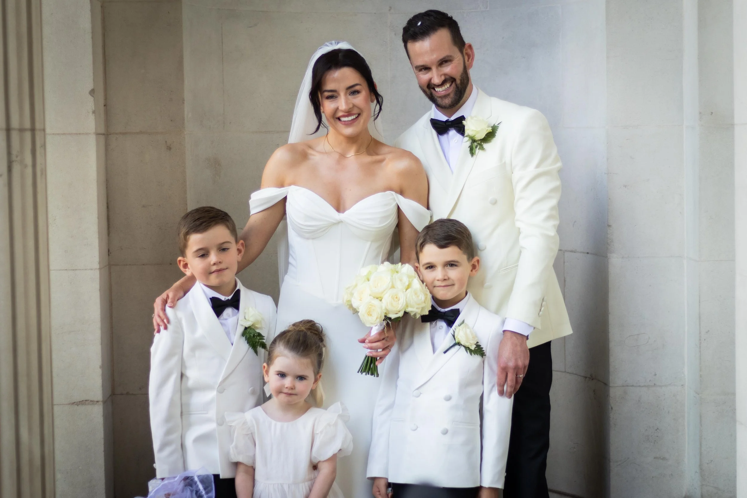 A wedding party photo of a bride and groom with three young boys and a young girl, all dressed in formal attire, standing indoors against a plain wall.
