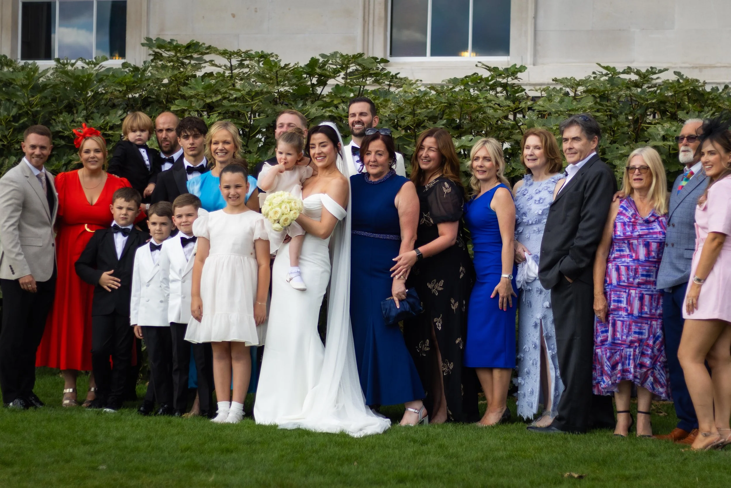 Group of people at a wedding, including bride in a white gown holding a bouquet, and groom in a tuxedo, standing outdoors on a lawn with greenery background.