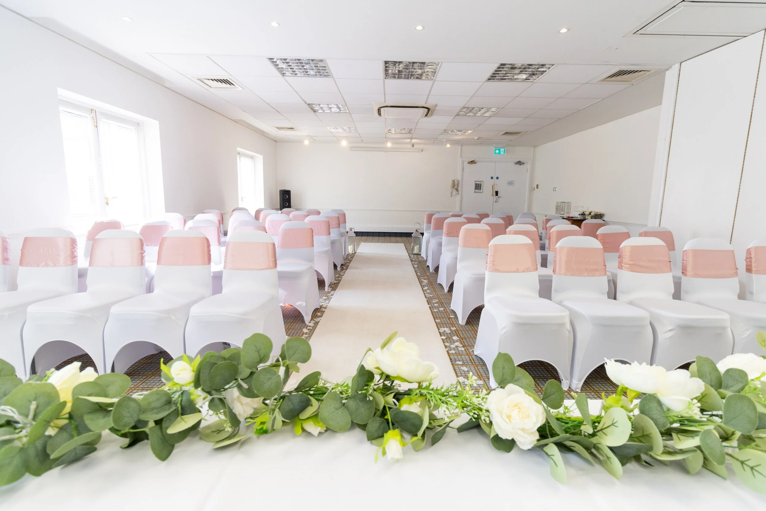 Empty wedding ceremony chairs with pink sash covers, facing a decorated aisle and floral arrangement at the front in a bright, white banquet hall.