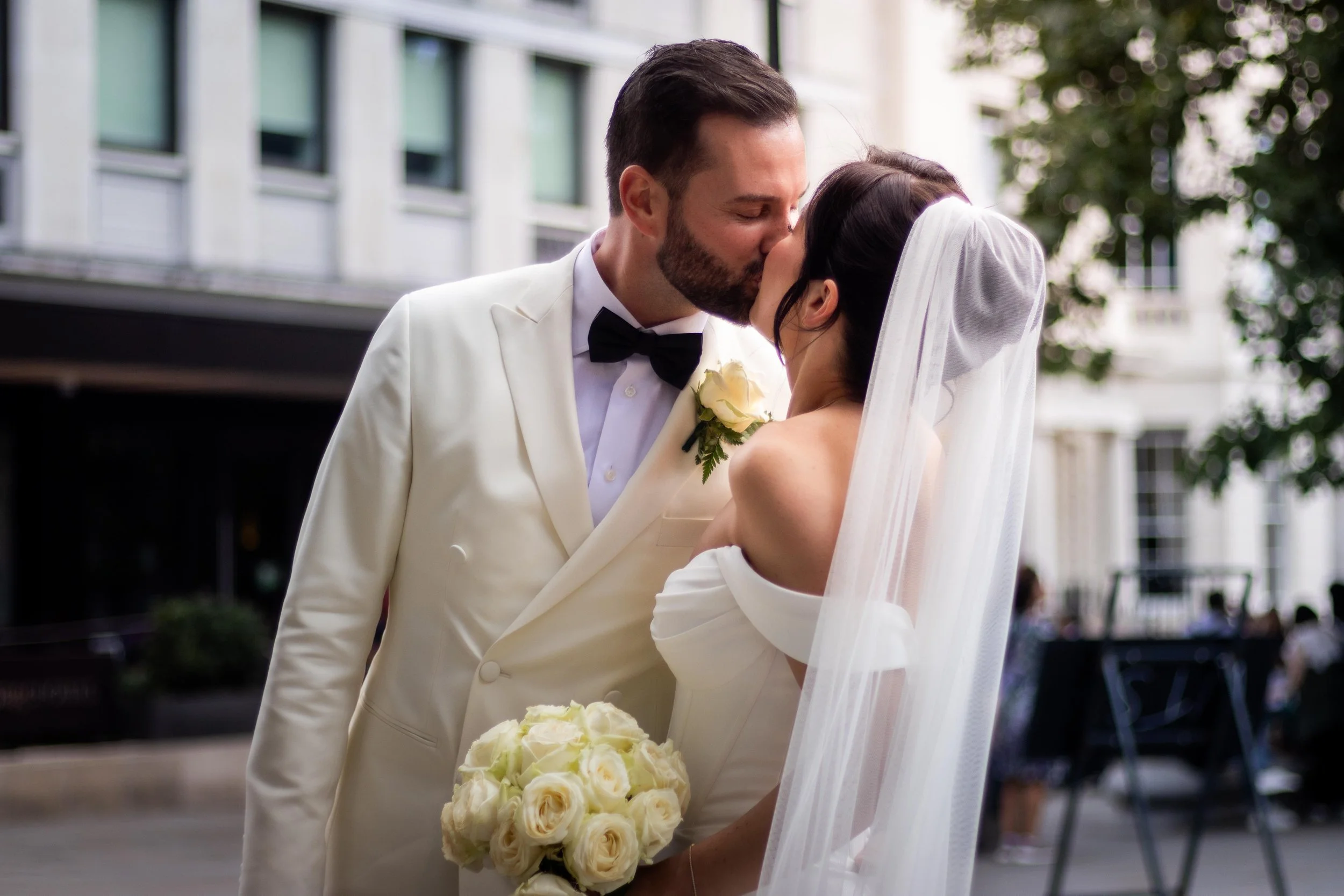 A newlywed couple sharing a kiss outdoors, with the groom in a white tuxedo and bow tie, and the bride in a white off-shoulder wedding dress holding a bouquet of white roses, and a white veil covering her hair.