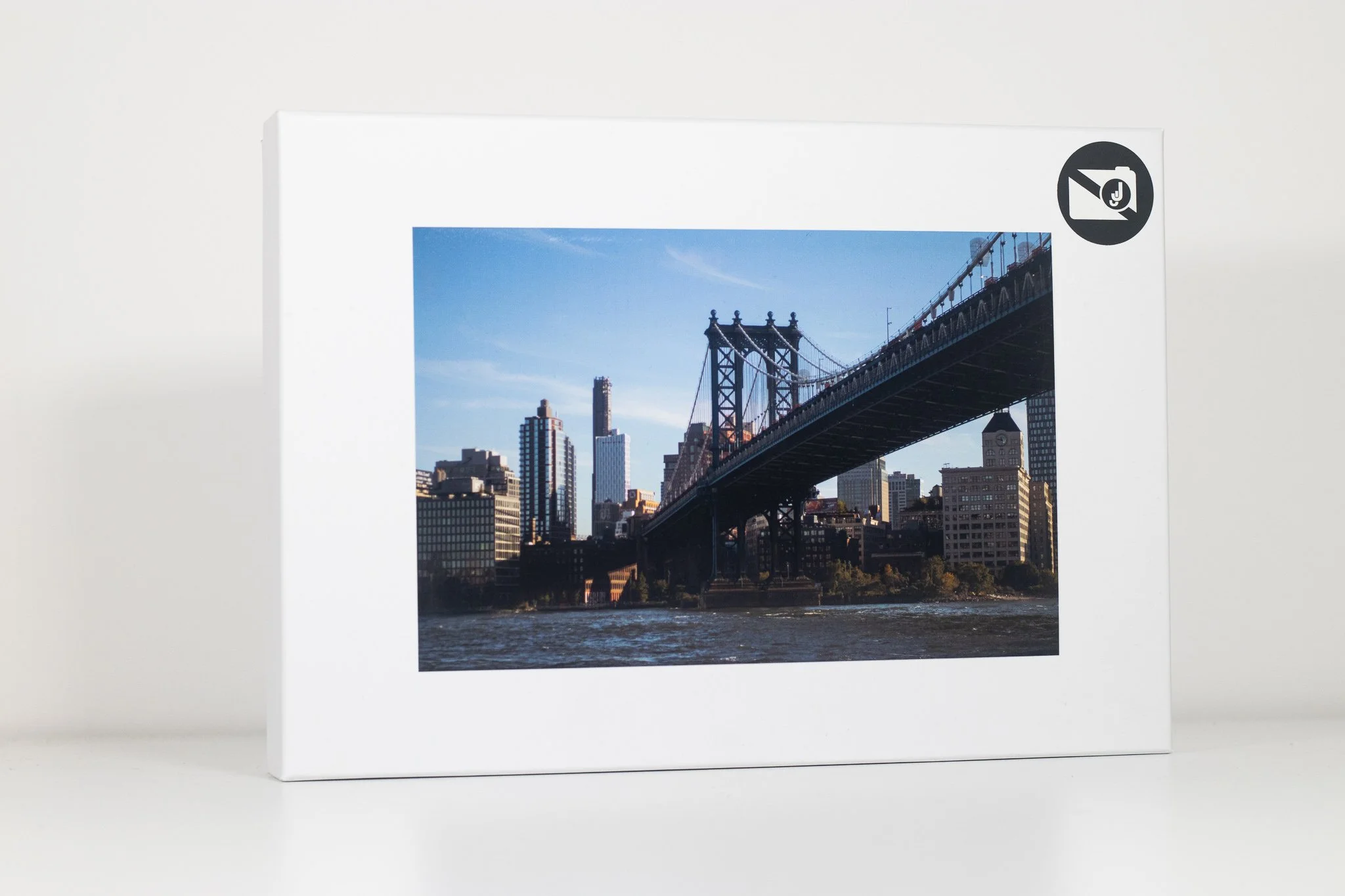 Framed photograph of a cityscape featuring a suspension bridge over a river, with skyscrapers in the background, under a clear blue sky.