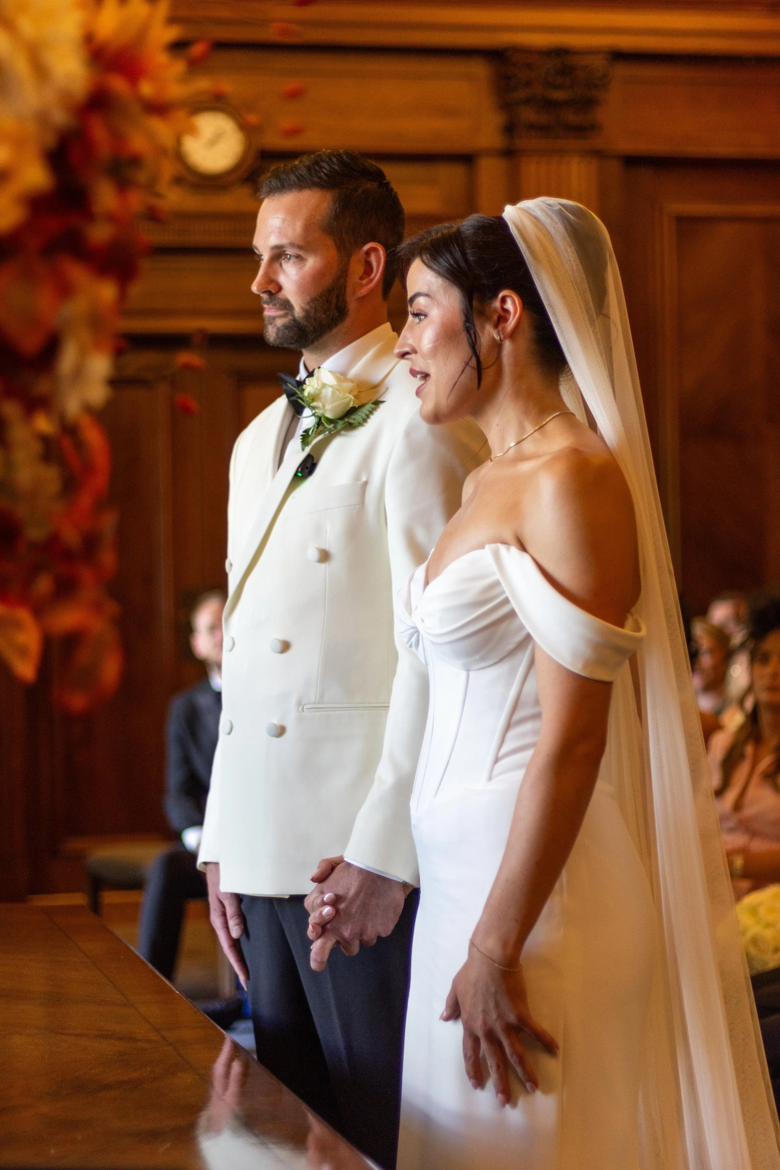 A bride and groom holding hands during their wedding ceremony, standing in front of a wood-paneled wall, with guests seated in the background.