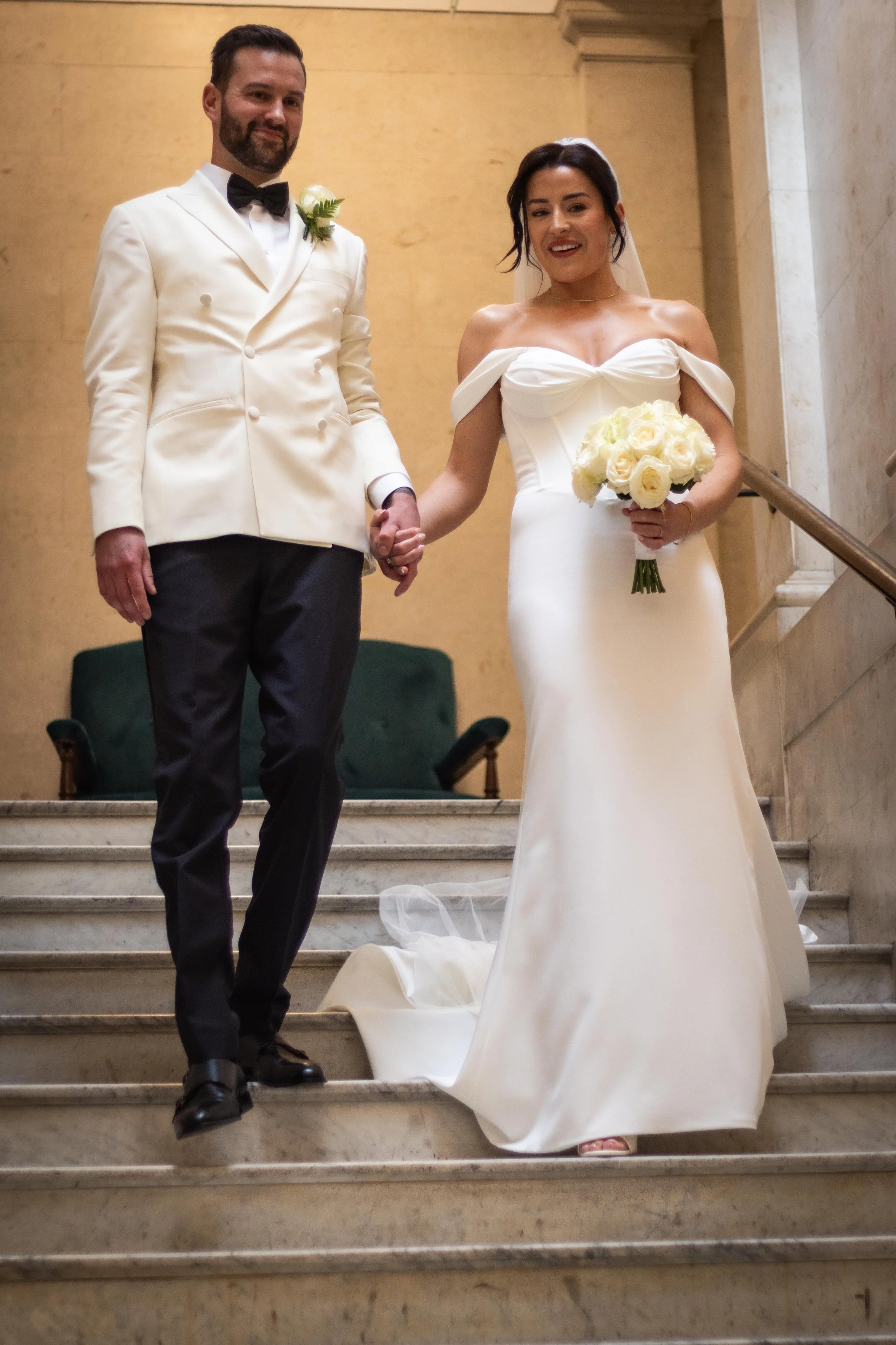 A bride and groom standing on stairs holding hands, the bride holding a bouquet of white roses, both smiling and dressed in wedding attire.
