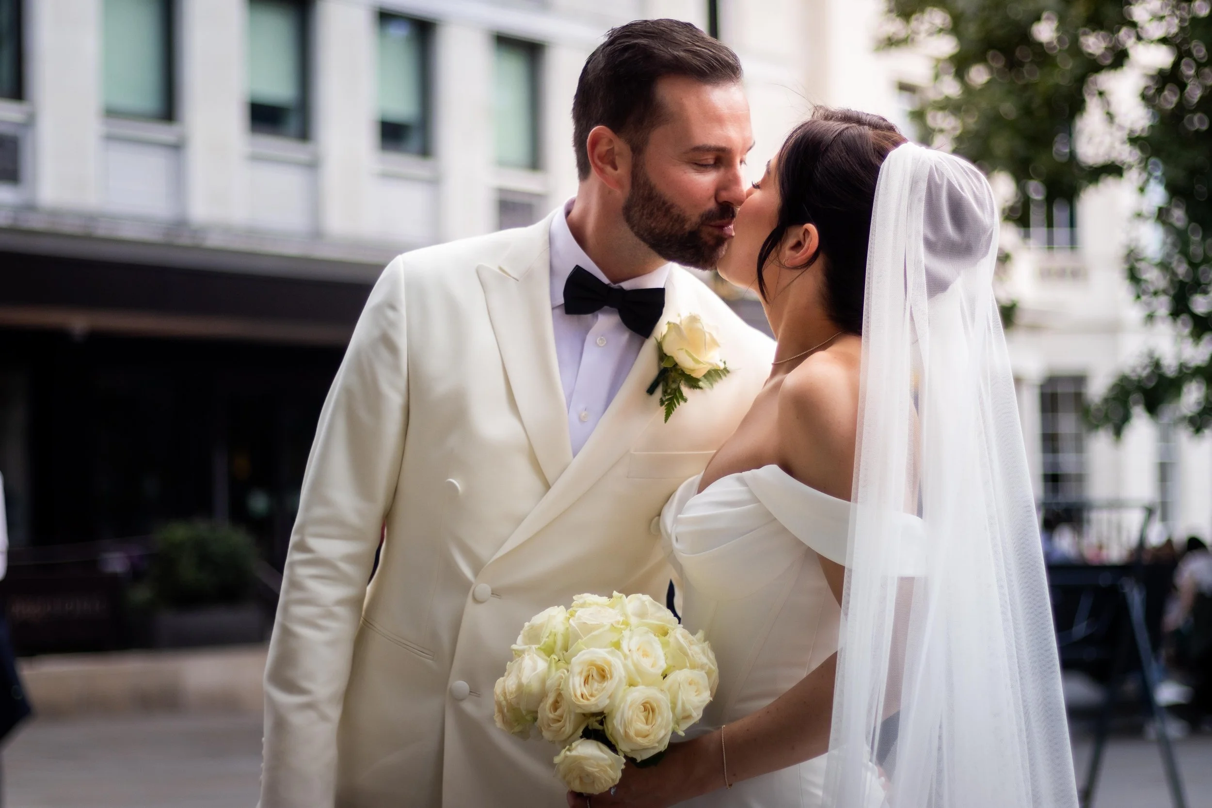 A newlywed couple sharing a kiss outdoors, with the groom in a white tuxedo and the bride in a white wedding dress holding a bouquet of white roses.