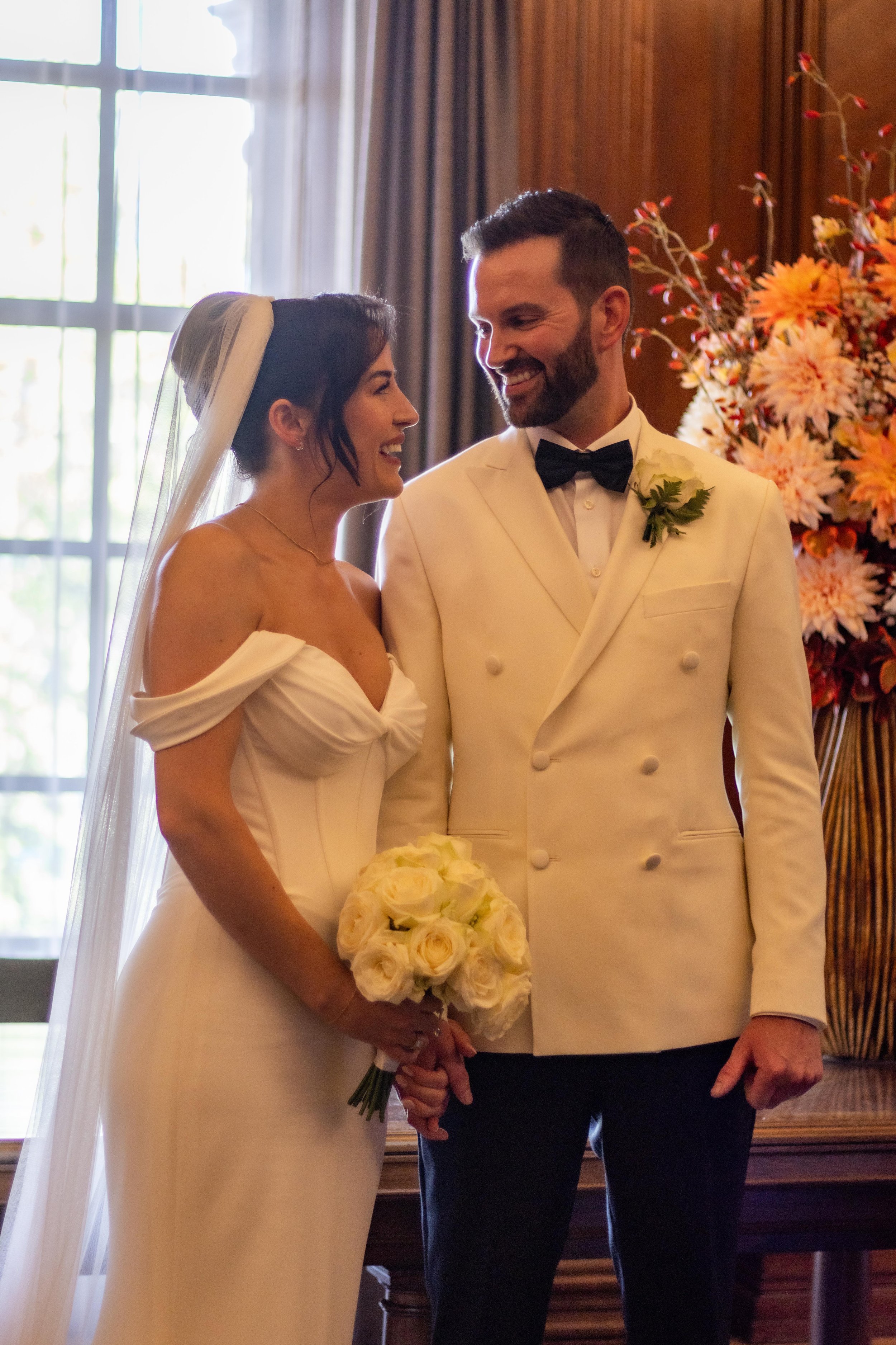 A bride and groom standing inside during their wedding ceremony, looking at each other and smiling, with flowers in the background.