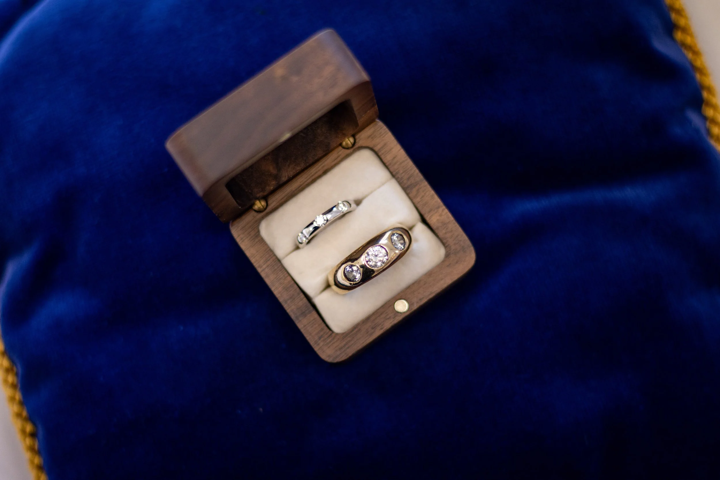 A wooden jewelry box containing three rings with diamonds, placed on a dark blue fabric background.