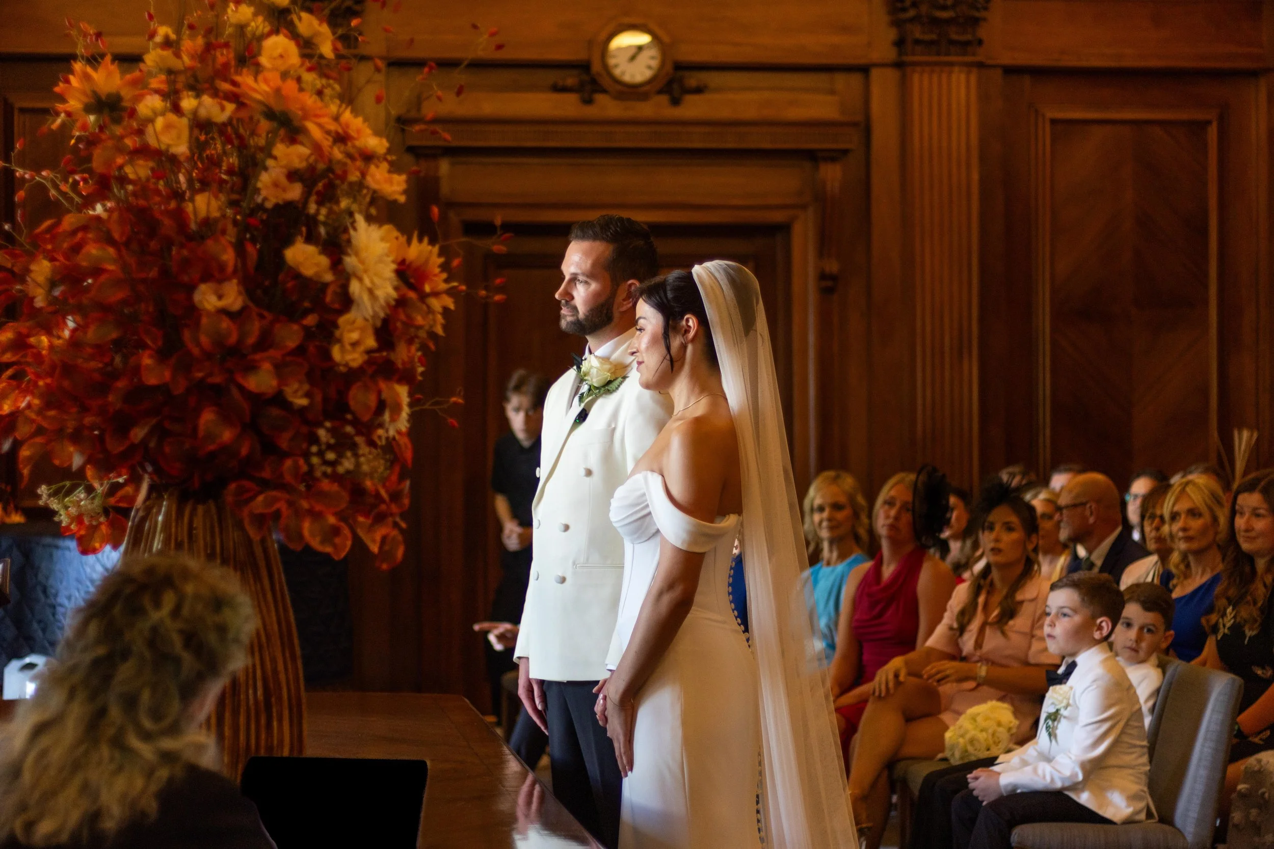 A bride and groom standing together during their wedding ceremony, facing forward in a wooden-paneled room, surrounded by seated guests.