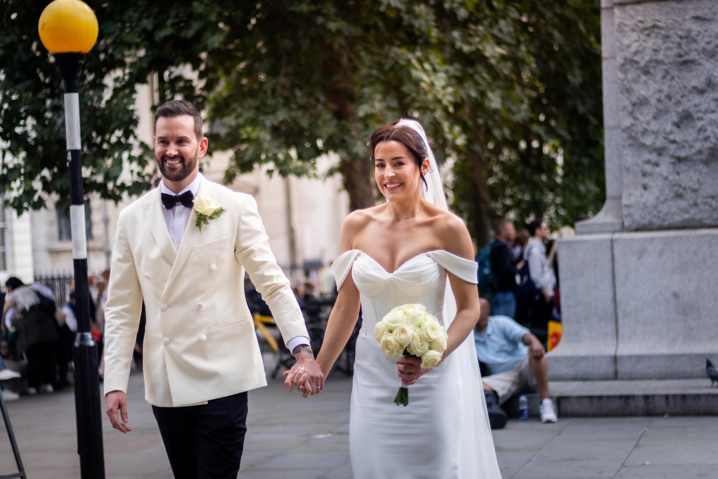 A smiling bride and groom walking hand-in-hand outdoors in a city, with the bride holding a bouquet of white roses, both dressed in wedding attire.
