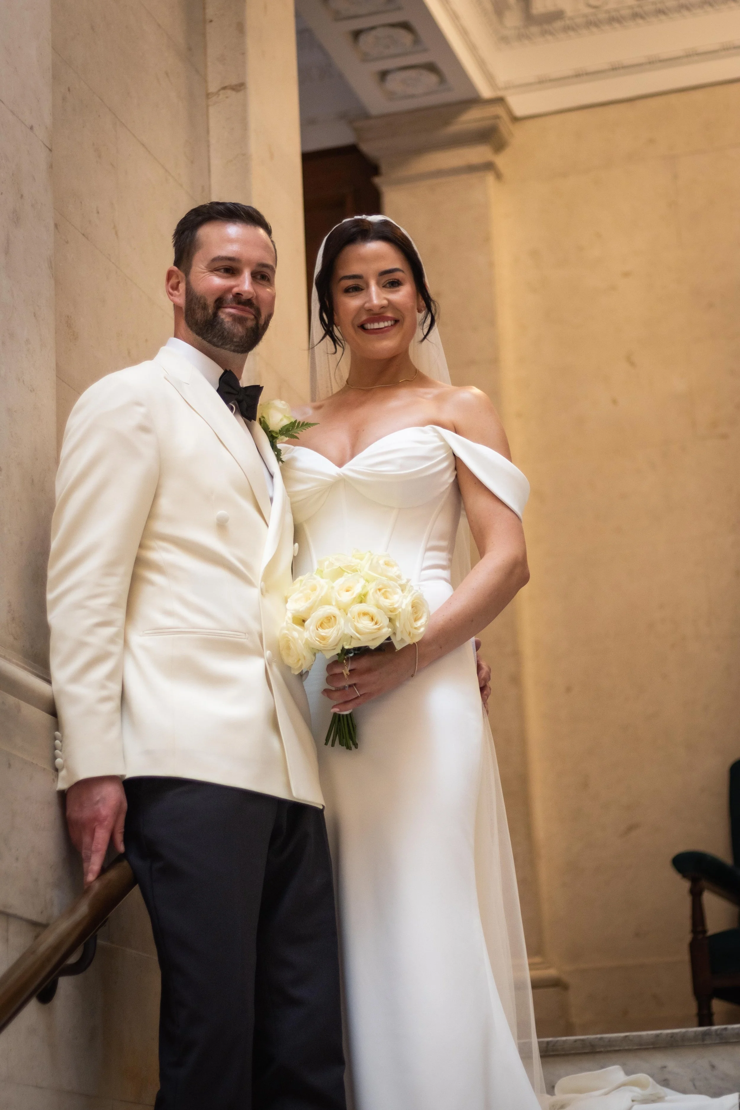 Bride and groom standing together in a wedding venue, smiling, with the bride holding a bouquet of white roses.