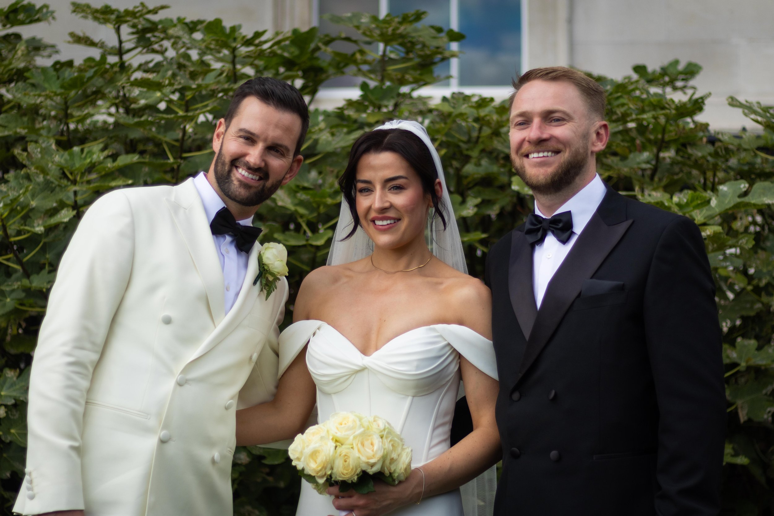 A bride in a white wedding dress holding a bouquet of white roses, flanked by two grooms in tuxedos, standing outdoors in front of greenery, smiling for the camera.