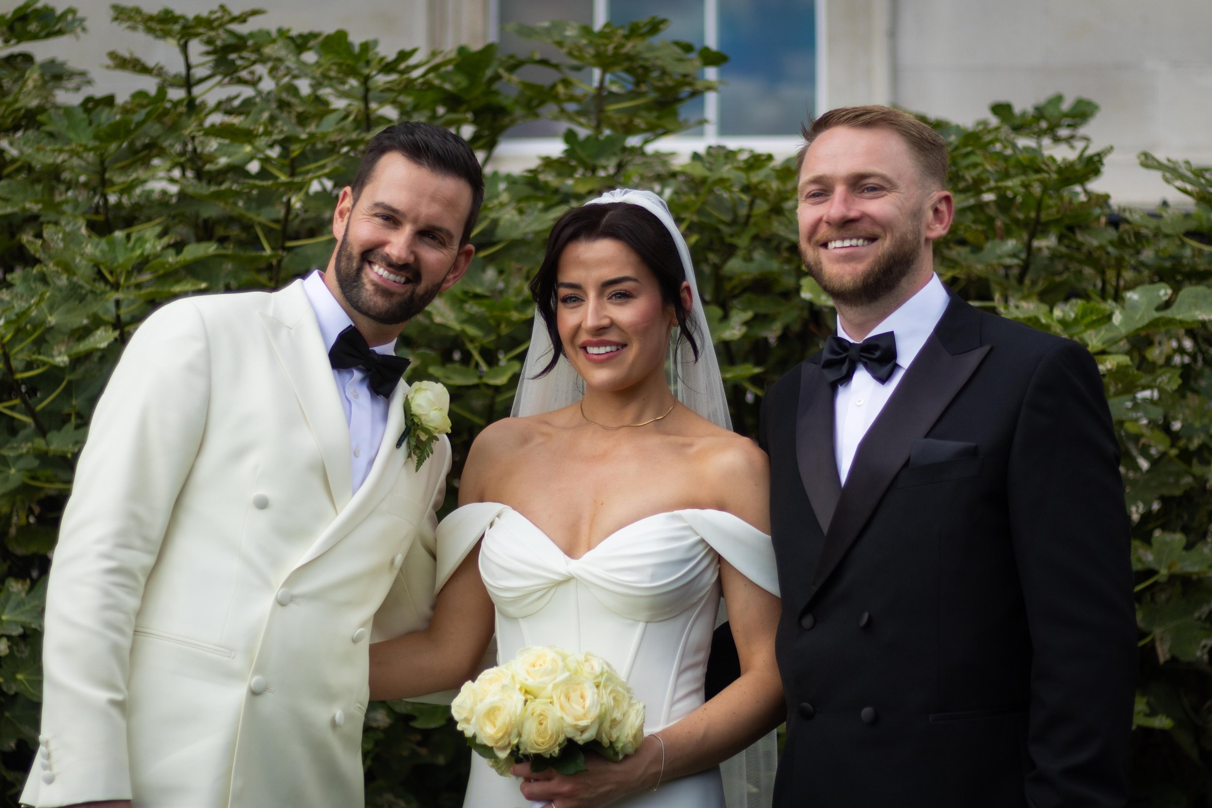 A bride in a white off-the-shoulder wedding dress holding a bouquet of white roses, flanked by two grooms in tuxedos, smiling outdoors with green foliage in the background.