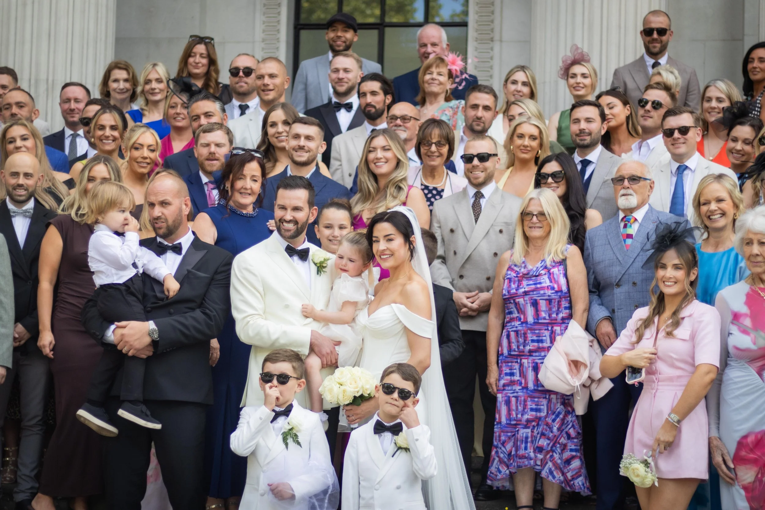 A large group of people gathered on the steps of a building, celebrating a wedding. The bride and groom are in the front center, surrounded by family and friends in colorful attire.