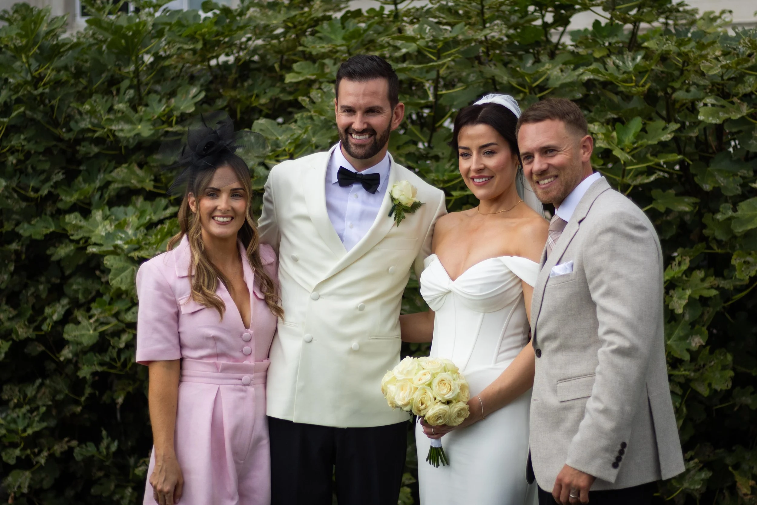 A group of five people at a wedding, including a bride holding a bouquet of white roses, a groom in a white tuxedo, two guests in colorful dresses, and a man in a light gray suit, standing outdoors in front of green foliage, all smiling.