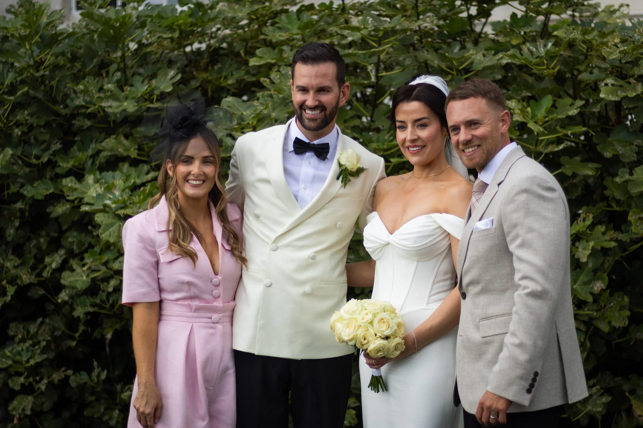 A group of four people dressed in wedding attire, standing outdoors in front of lush green foliage, smiling for a photo. The group includes a bride holding a bouquet of white roses, a groom in a white tuxedo with a black bow tie, a woman in a pink ou