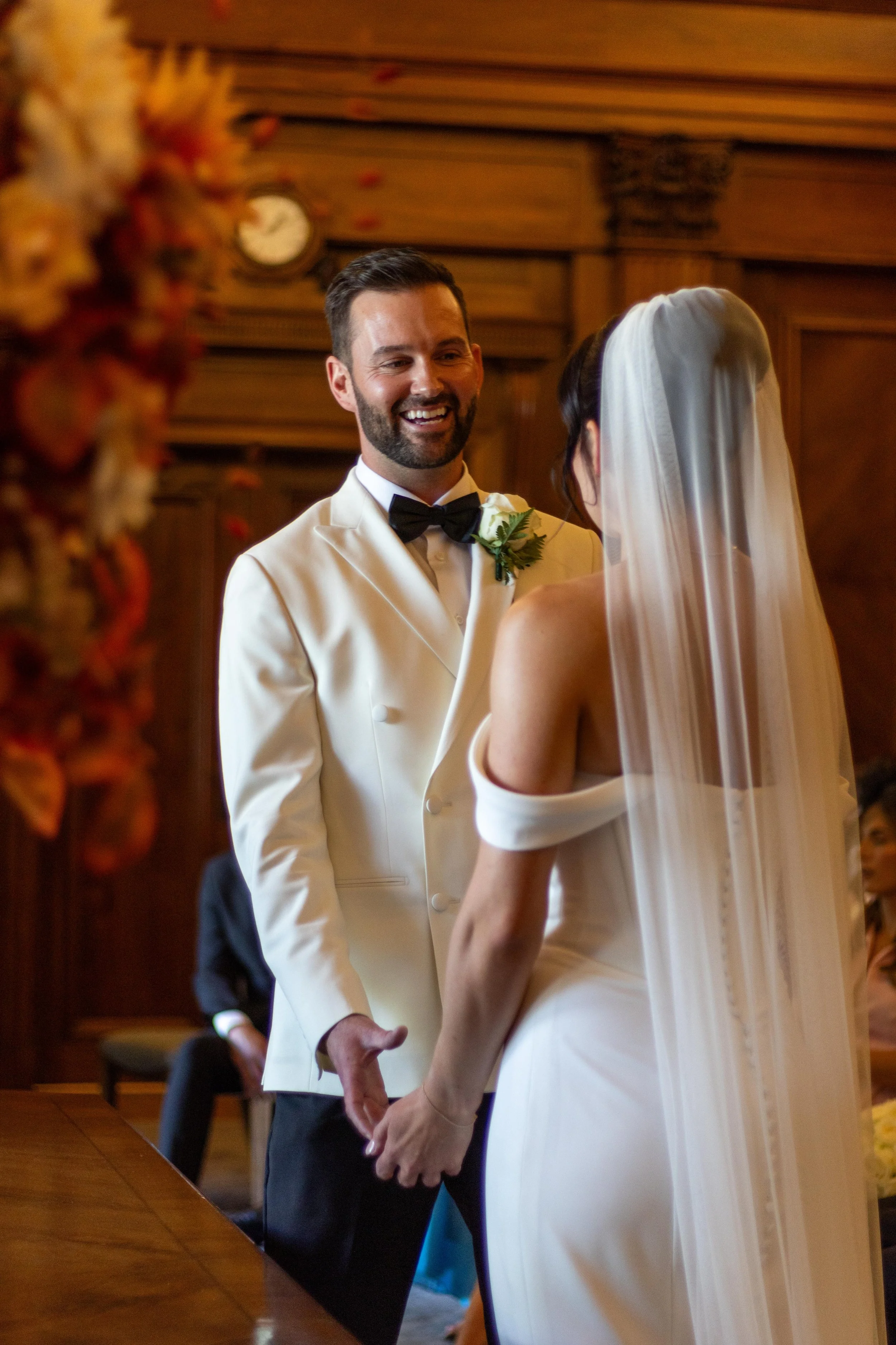A wedding ceremony with a groom in a white tuxedo and a bride in a white wedding dress, holding hands and smiling at each other inside a wooden hall.