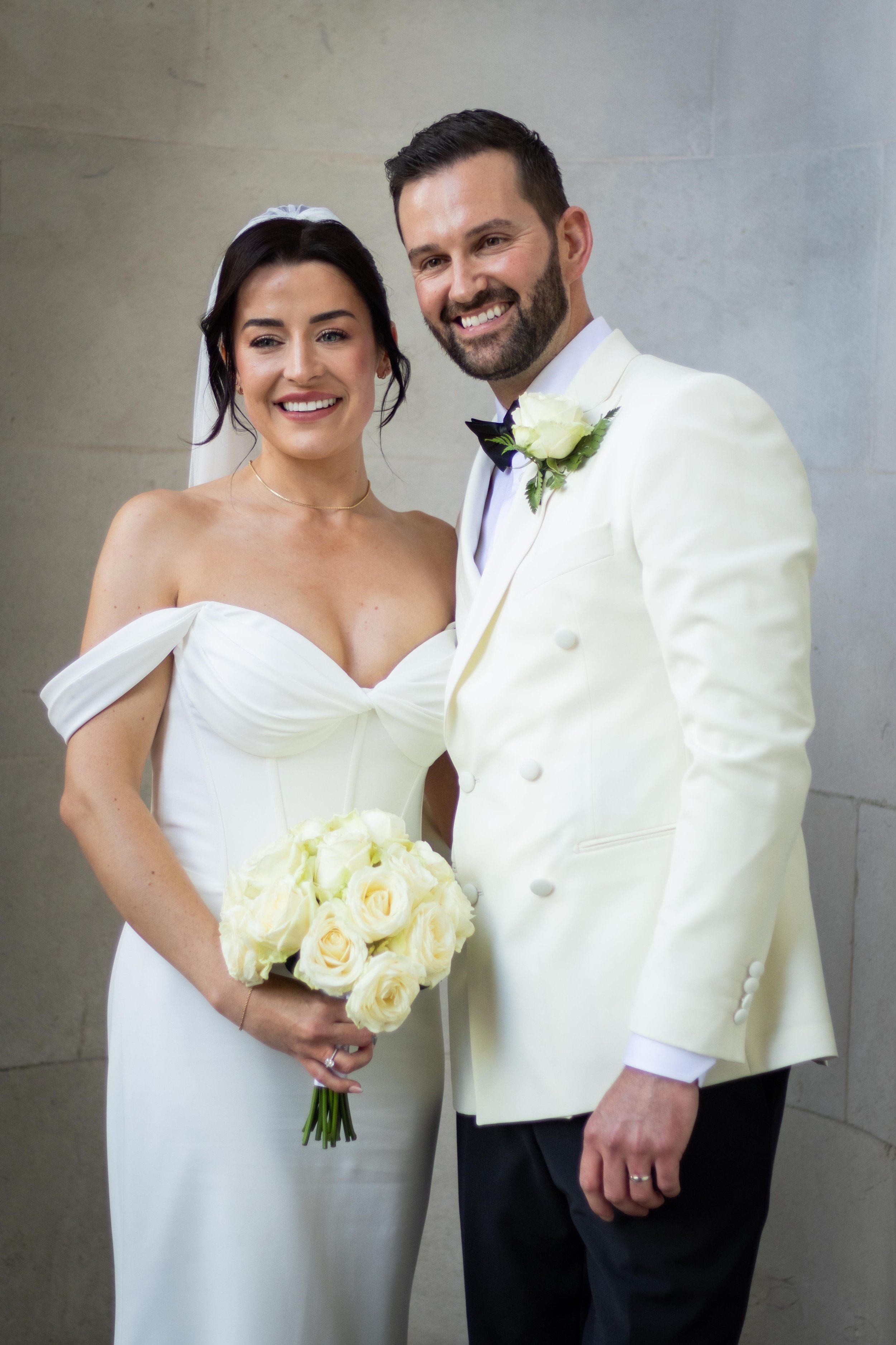 A bride and groom smiling at their wedding, standing close together in formal attire, with the bride holding a bouquet of white roses.