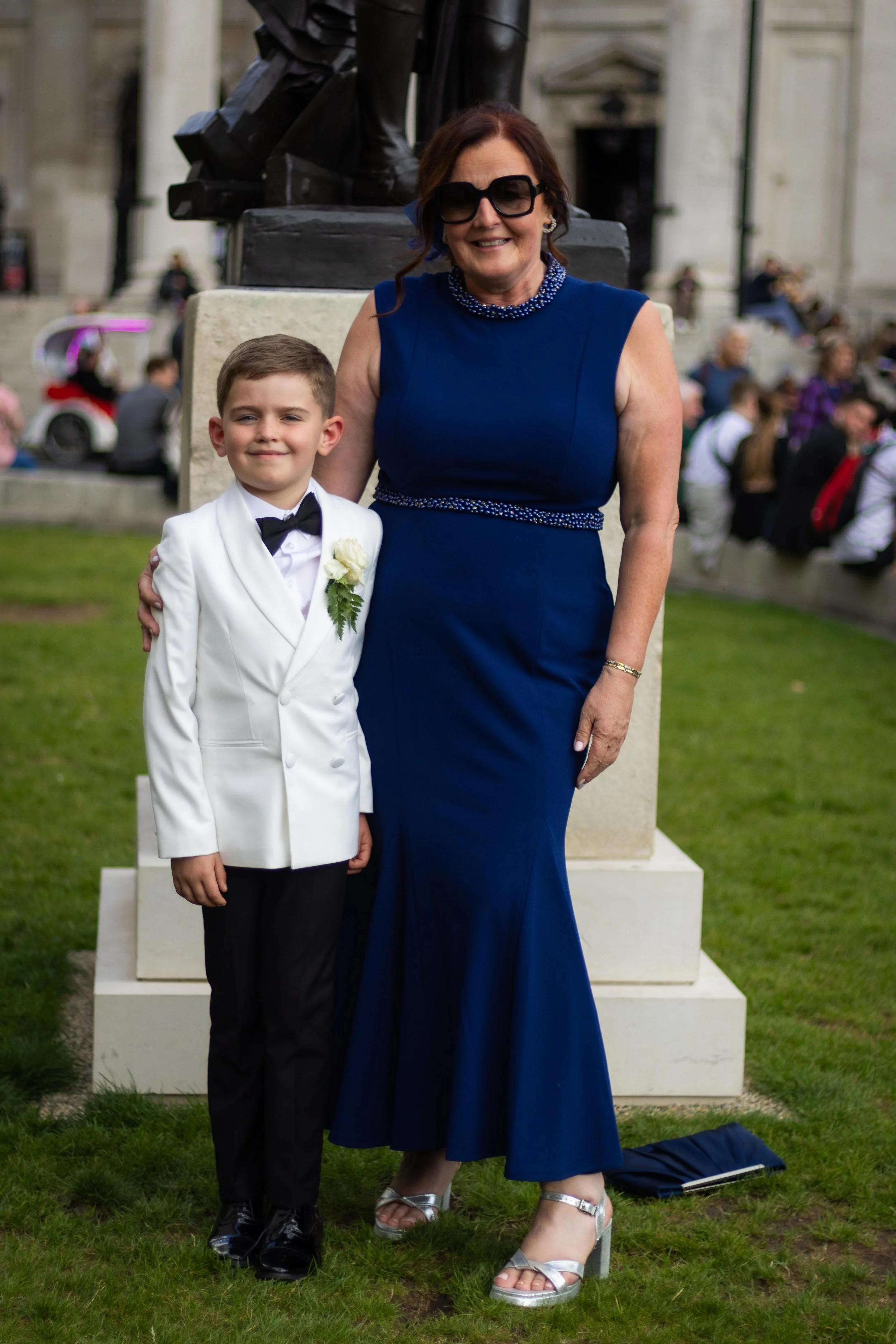 A boy in a white tuxedo with a bow tie standing next to a woman in a blue dress at a formal outdoor event.
