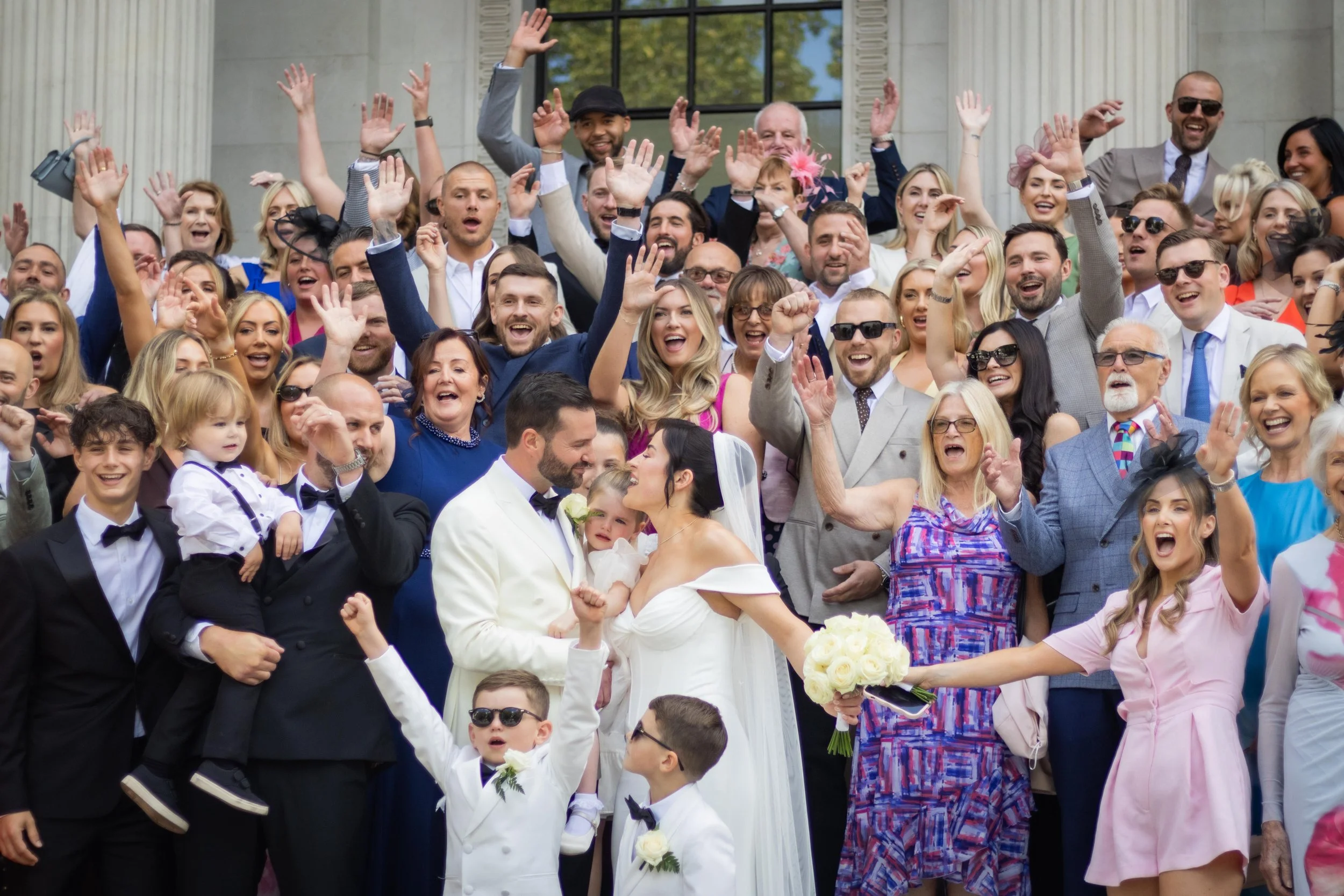 A large group of people celebrating a wedding outside a building, with a bride and groom in the center, all smiling and cheering.