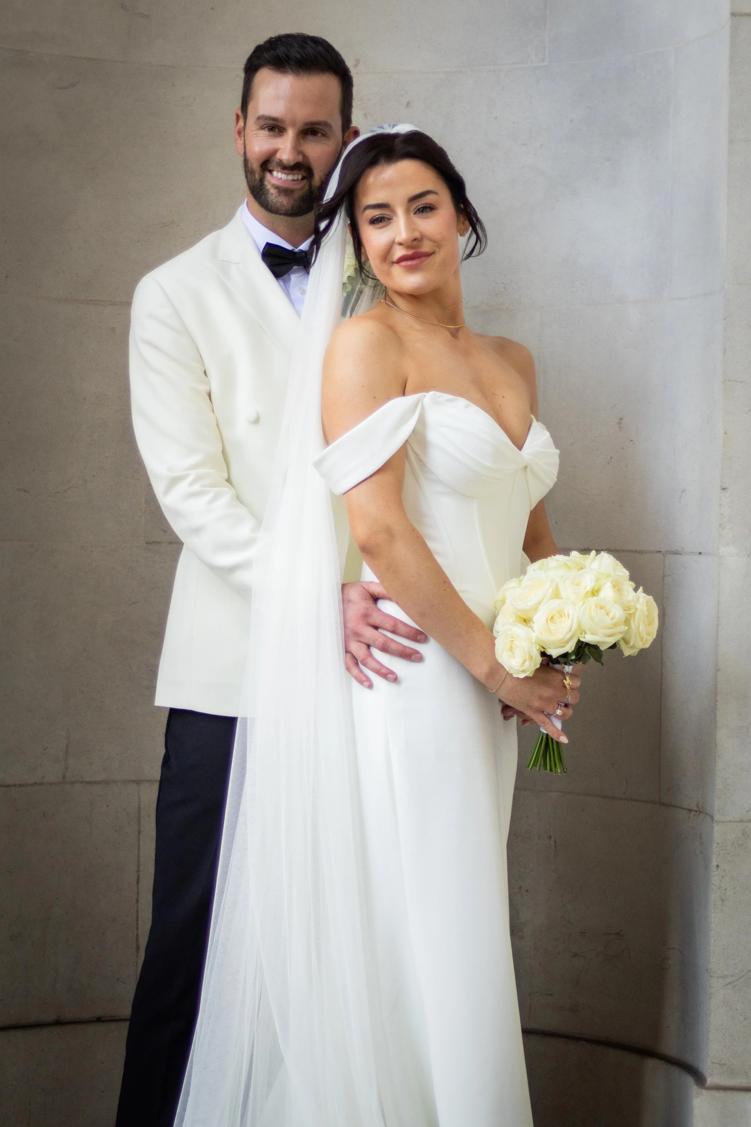 A bride and groom pose together at their wedding, with the groom standing behind the bride and holding her waist. The bride is holding a bouquet of white roses and is dressed in a white off-the-shoulder gown. The groom wears a white tuxedo jacket wit