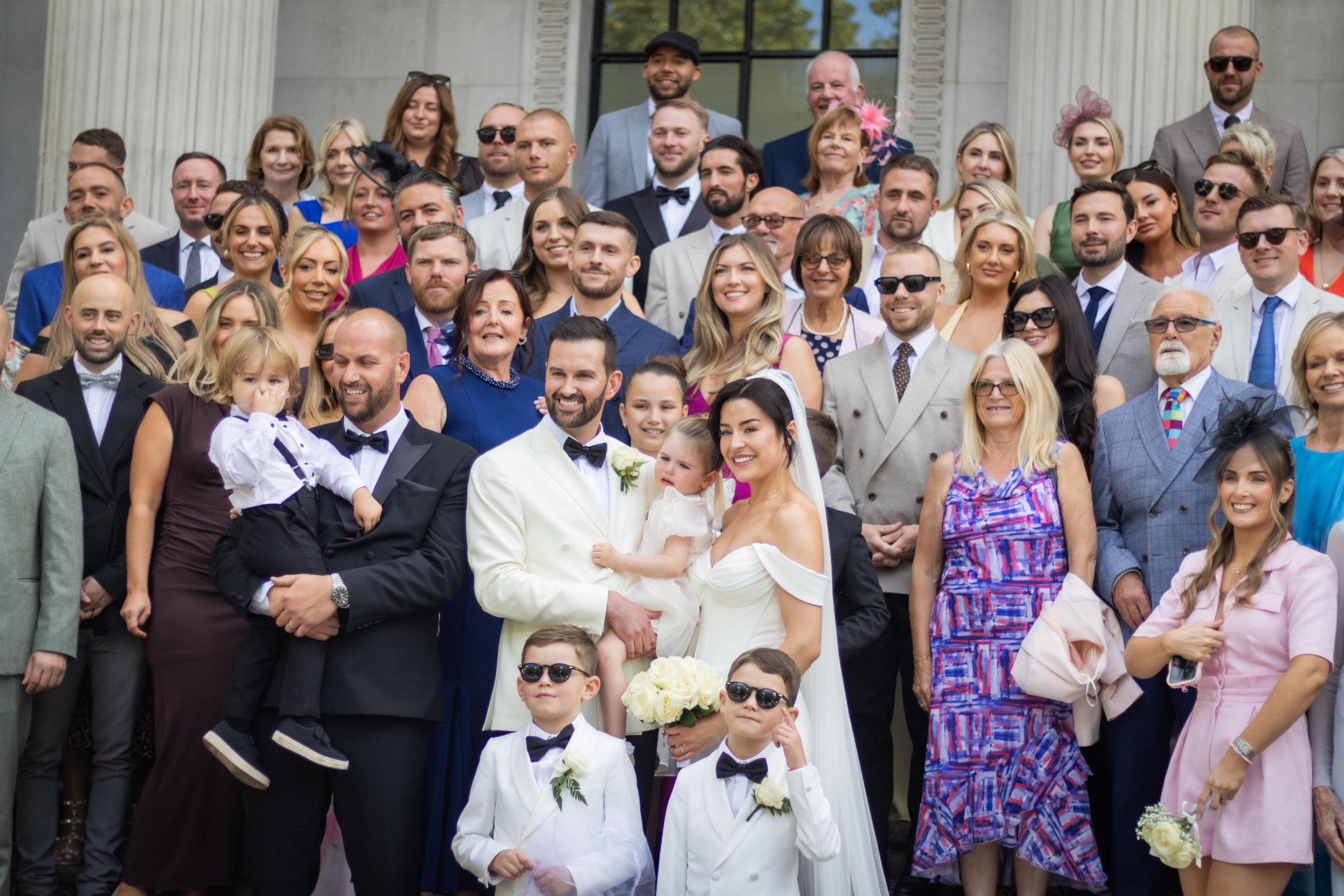 Group photo of wedding guests outside a building featuring a bride and groom in the center, surrounded by smiling friends and family in formal attire, with some wearing sunglasses and colorful dresses.