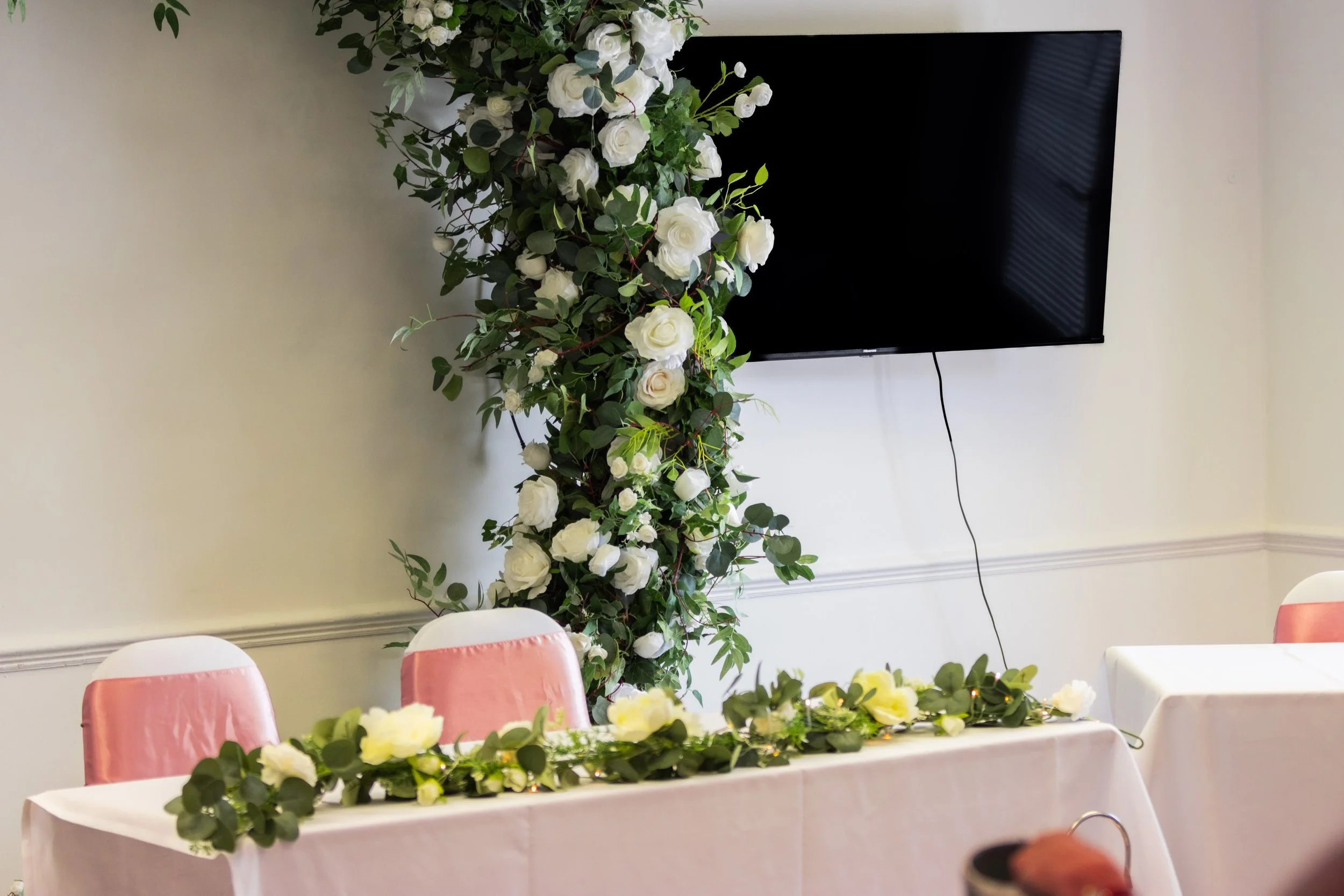 Table decorated with white flowers and greenery, with pink and white chairs in a room with a white wall, a large floral arrangement with white roses, and a television mounted on the wall.
