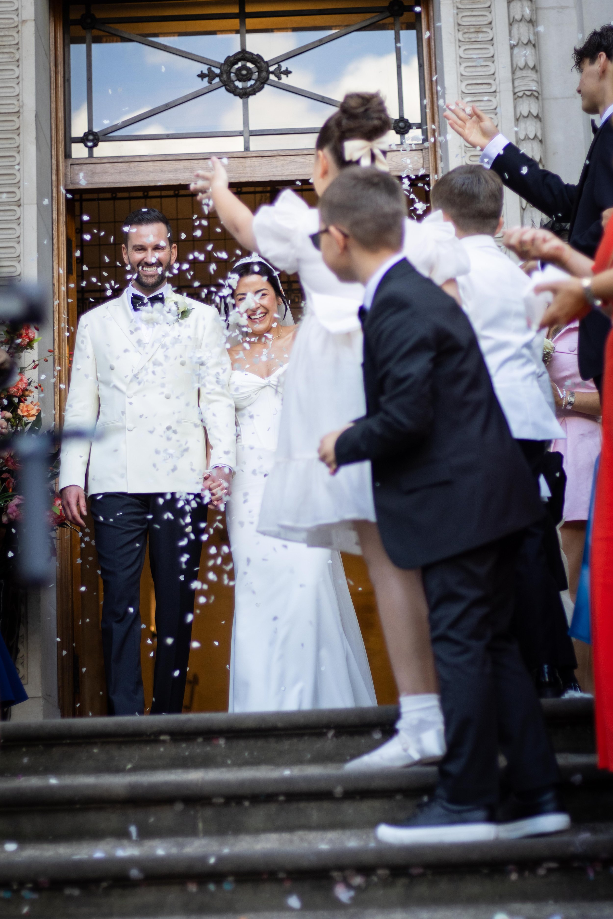 A newlywed couple in wedding attire exiting a building while friends celebrate with confetti. The bride and groom are smiling and holding hands, surrounded by guests dressed formally.