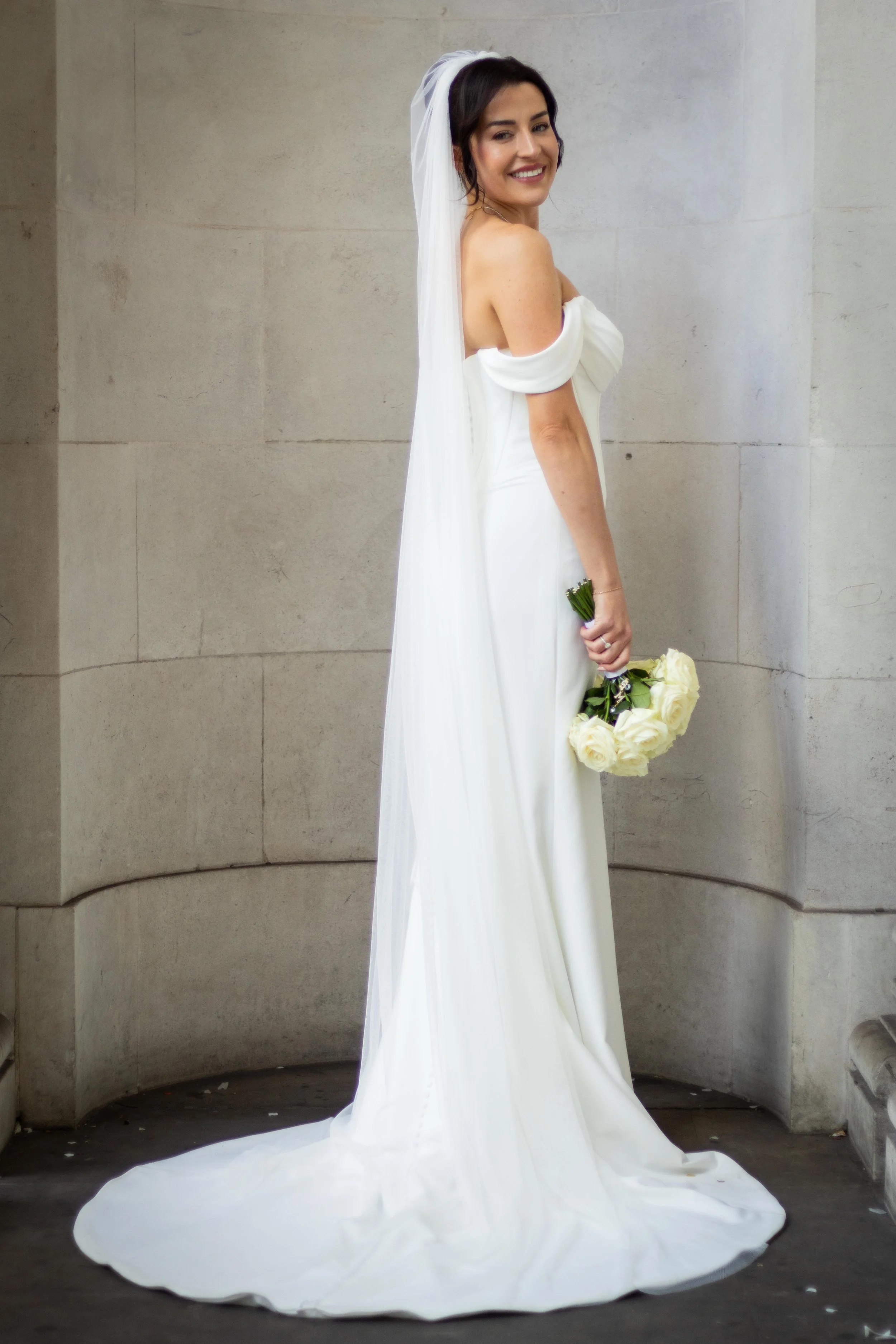 A smiling bride in a white wedding dress with off-shoulder sleeves, holding a bouquet of white roses, standing against a stone wall.