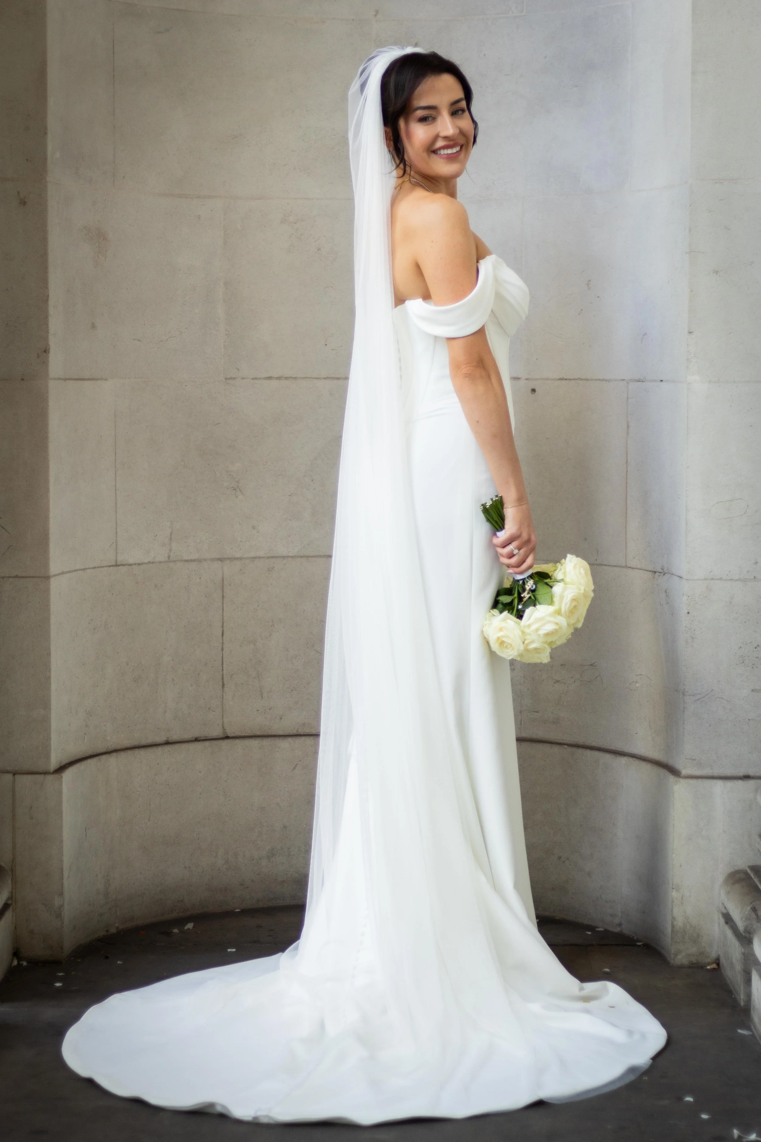 Brunette woman in a white wedding dress holding a bouquet of white roses, standing against a stone wall.