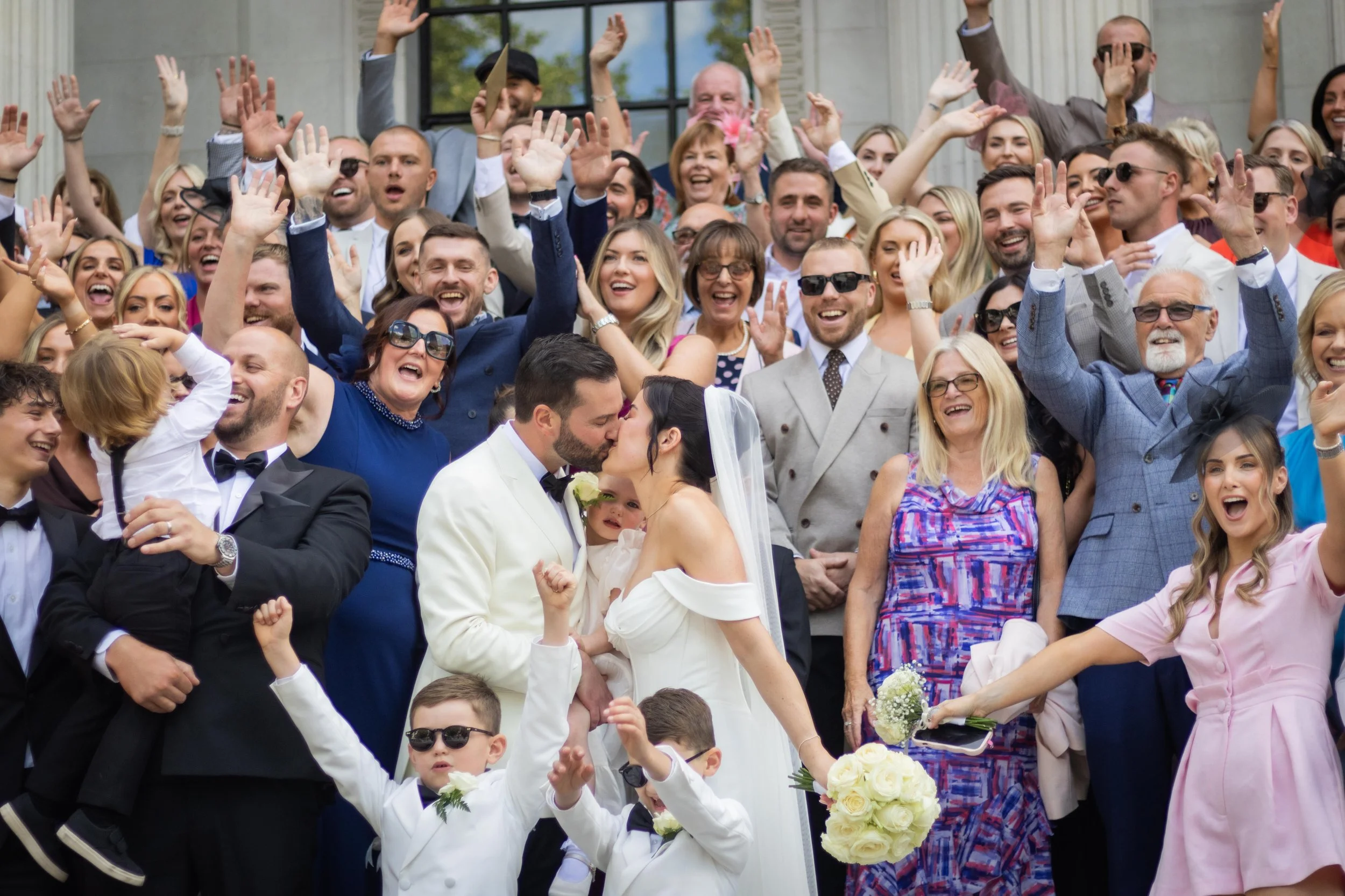 A large group of wedding guests celebrating outside, with many raising their hands and smiling. The bride and groom are in the center, sharing a kiss, with the bride holding a bouquet of white roses. Children, women, and men in formal attire fill the